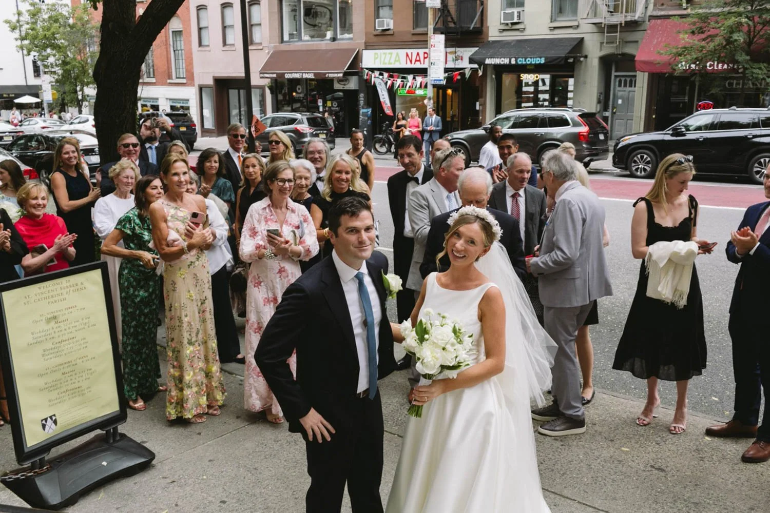 A bride and groom smiling in front of a crowd of friends and family outside a church during a wedding celebration.