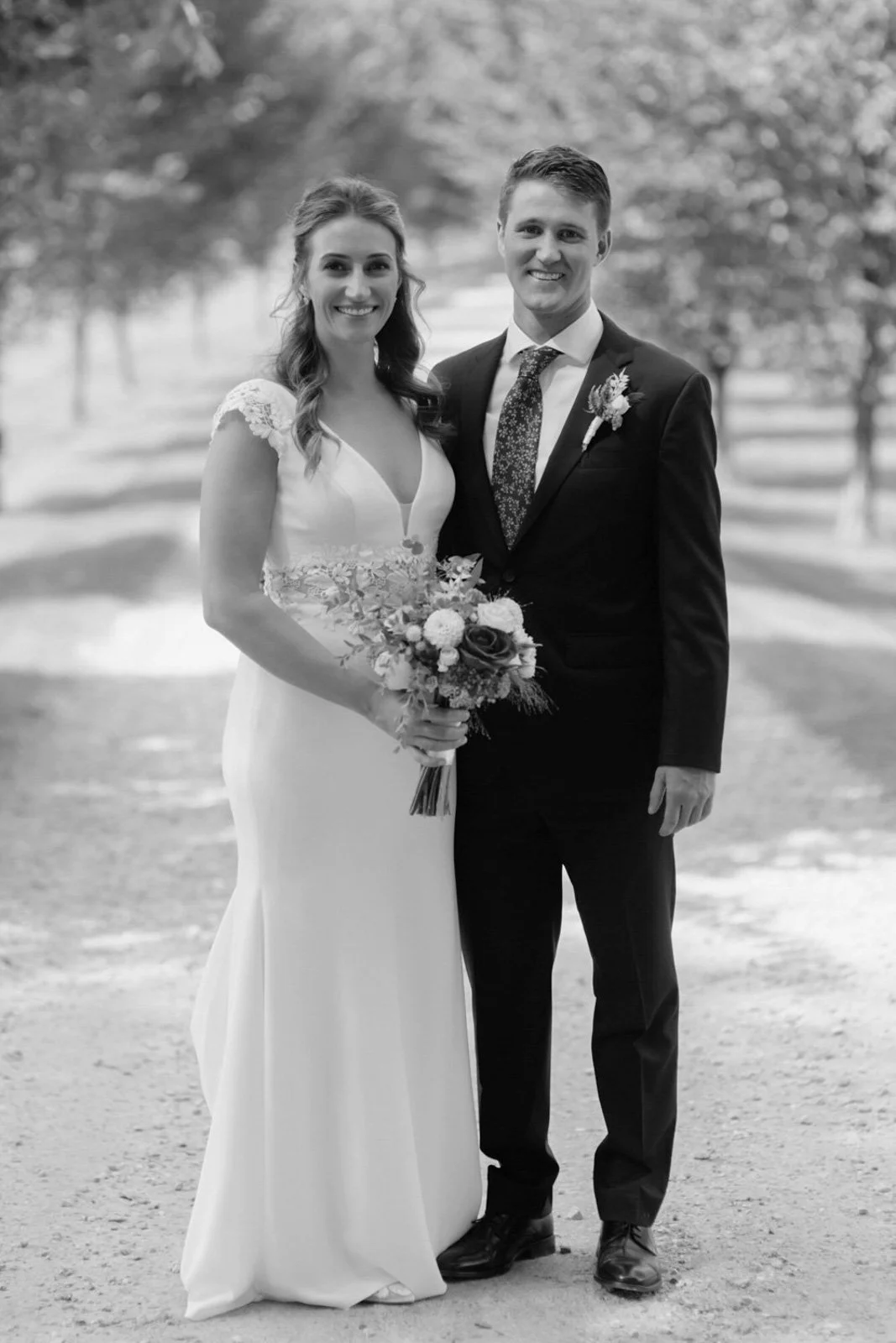 Black and white photo of a bride and groom standing outdoors with trees in the background. The bride is holding a bouquet and wearing a wedding gown with lace details, and the groom is in a dark suit with a floral tie.