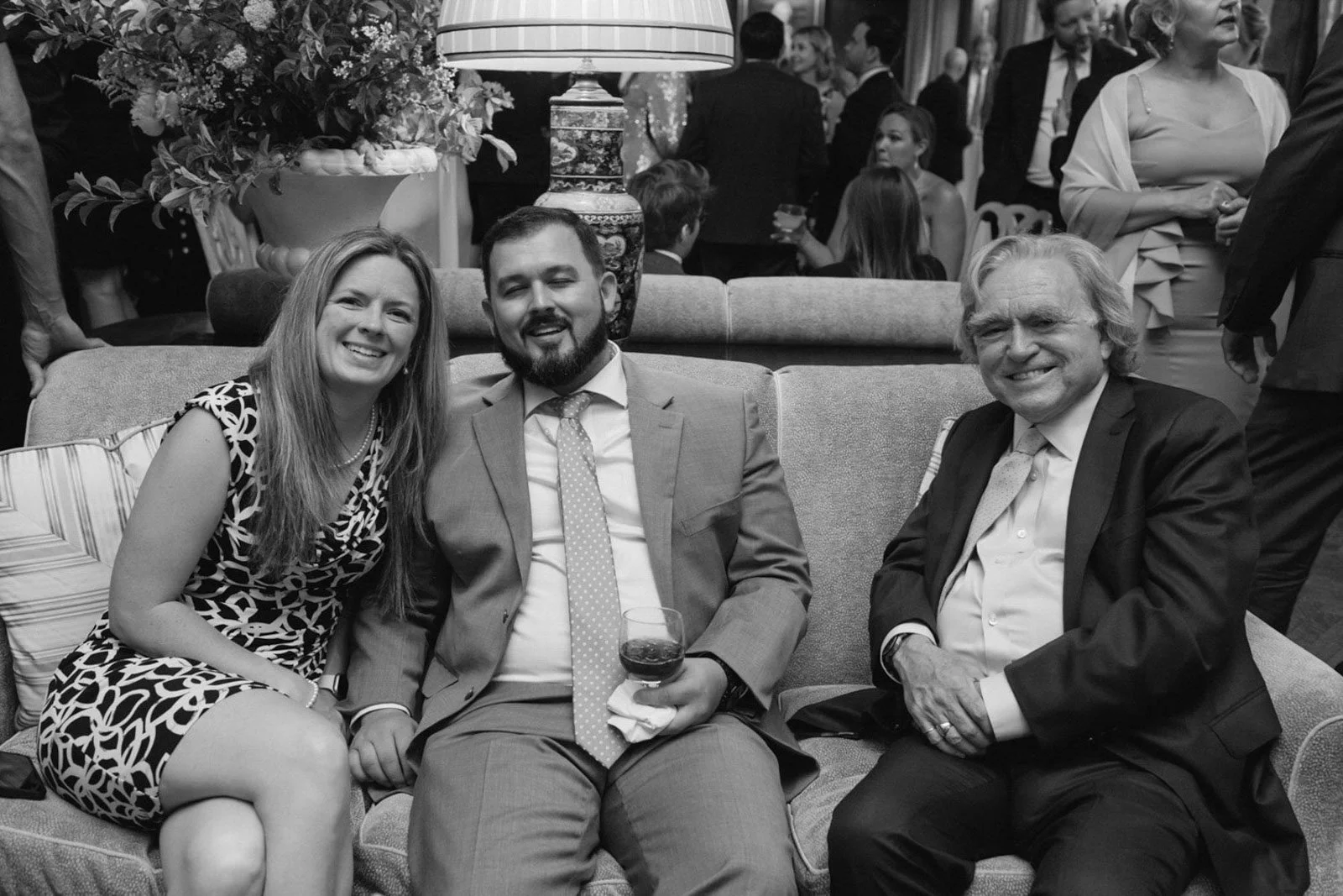 Three people sitting on a couch at a social gathering, with a woman in a patterned dress on the left, a man in a suit with a wine glass in the middle, and an older man in a suit on the right, smiling for the photo.