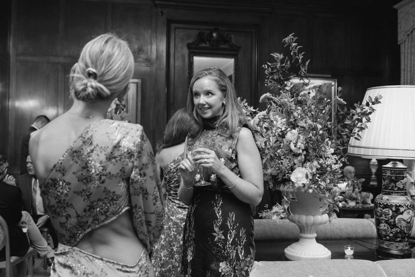Black and white photo of two women at a social gathering, one holding a glass of wine, in a room with wood-paneled walls, floral arrangements, and decorative lamps.