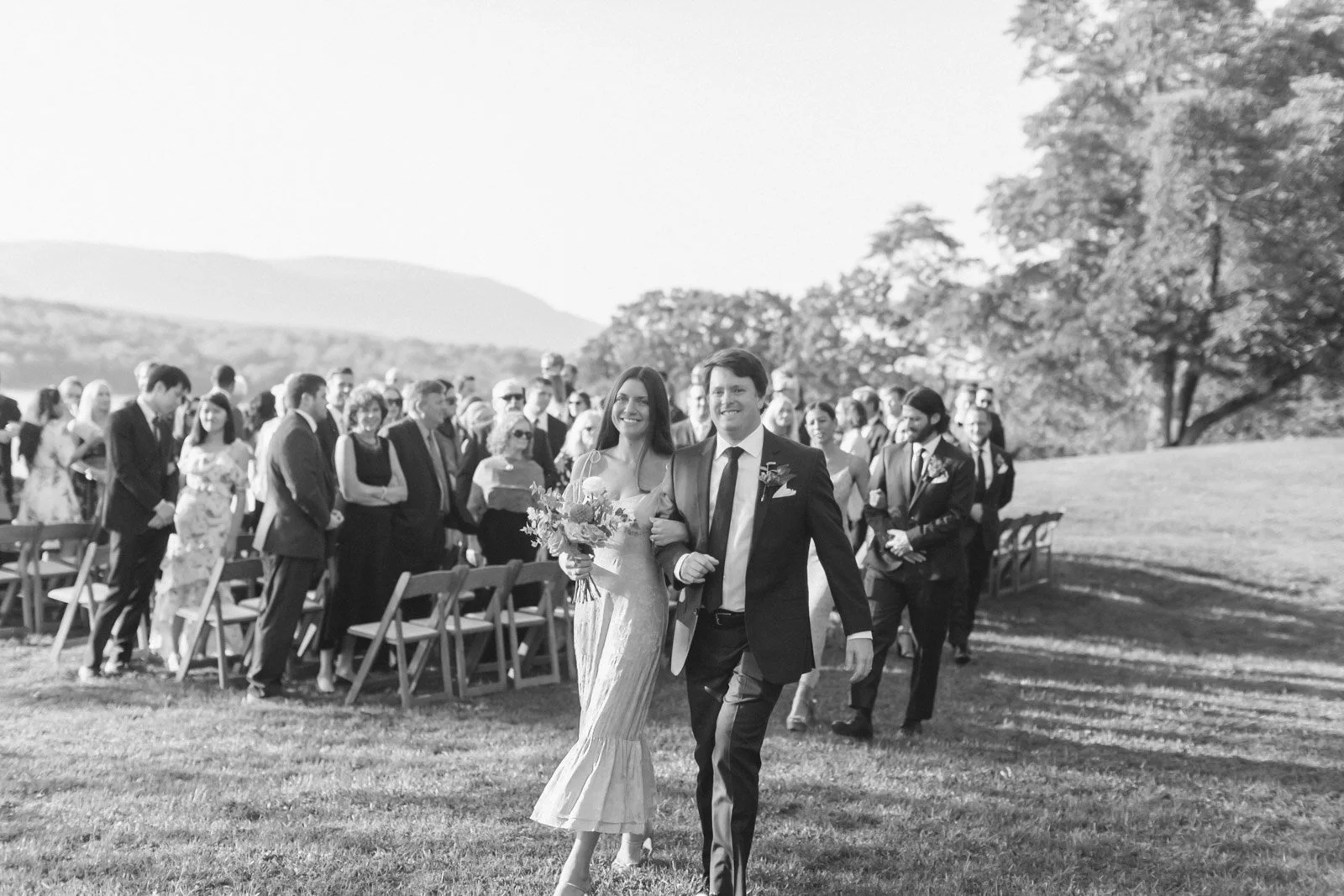 A black-and-white photo of a wedding scene outdoors with a bride and groom walking arm-in-arm in the foreground, smiling, with guests in the background, some seated and some standing, on a grassy area with trees and hills in the distance.