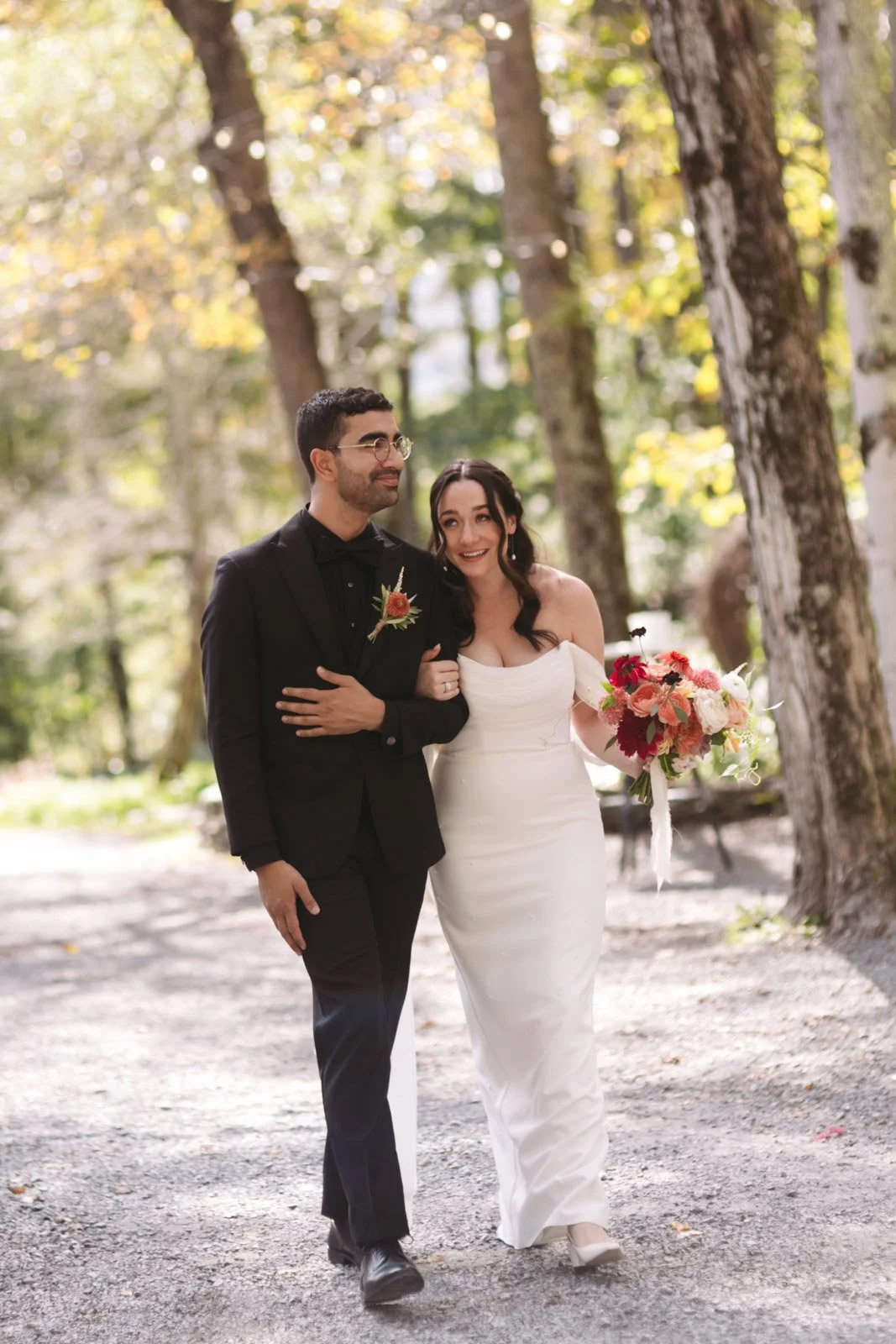 A newlywed couple walking arm in arm in a wooded park, with trees in the background. The bride is holding a bouquet of flowers and is smiling, wearing a white off-the-shoulder wedding dress. The groom is dressed in a black suit with a boutonnière.