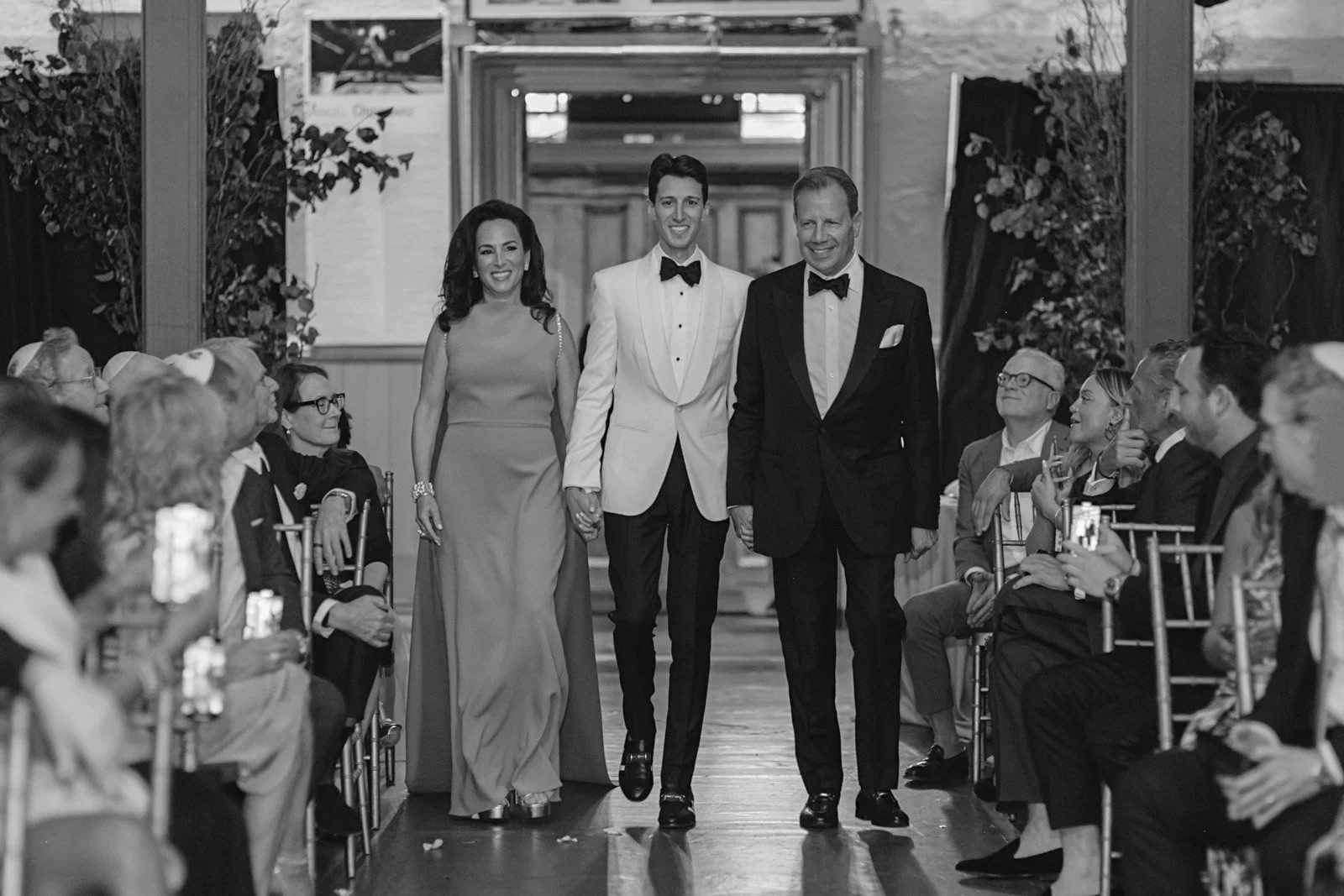 Black and white photo of a wedding ceremony, with three people walking down the aisle holding hands; the couple in the middle wears a tuxedo and the woman on the left wears a gown. Guests seated on both sides watch and take photos.