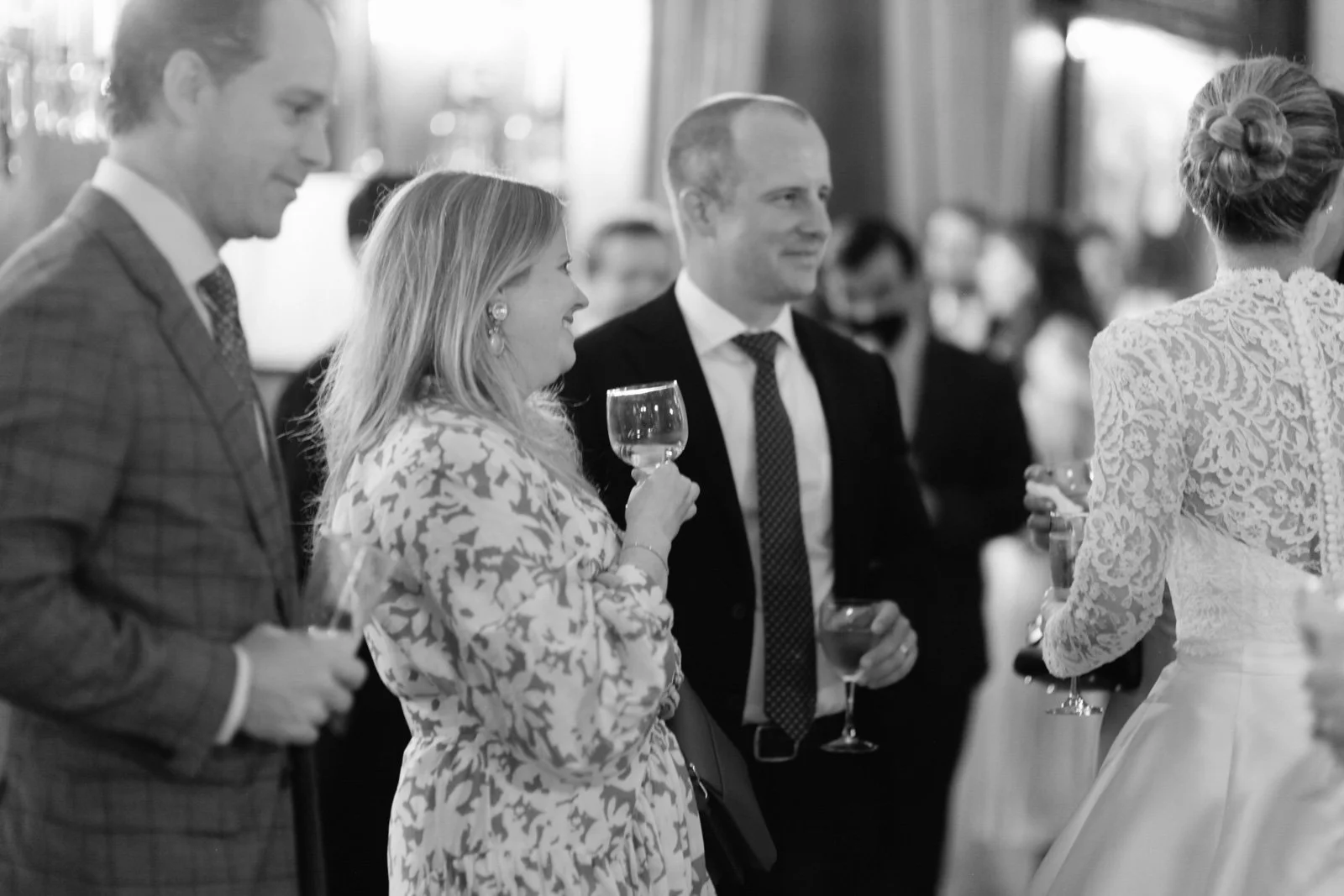 People socializing at a formal event, holding wine glasses, with a woman in a lace dress and others dressed in suits and formal attire.