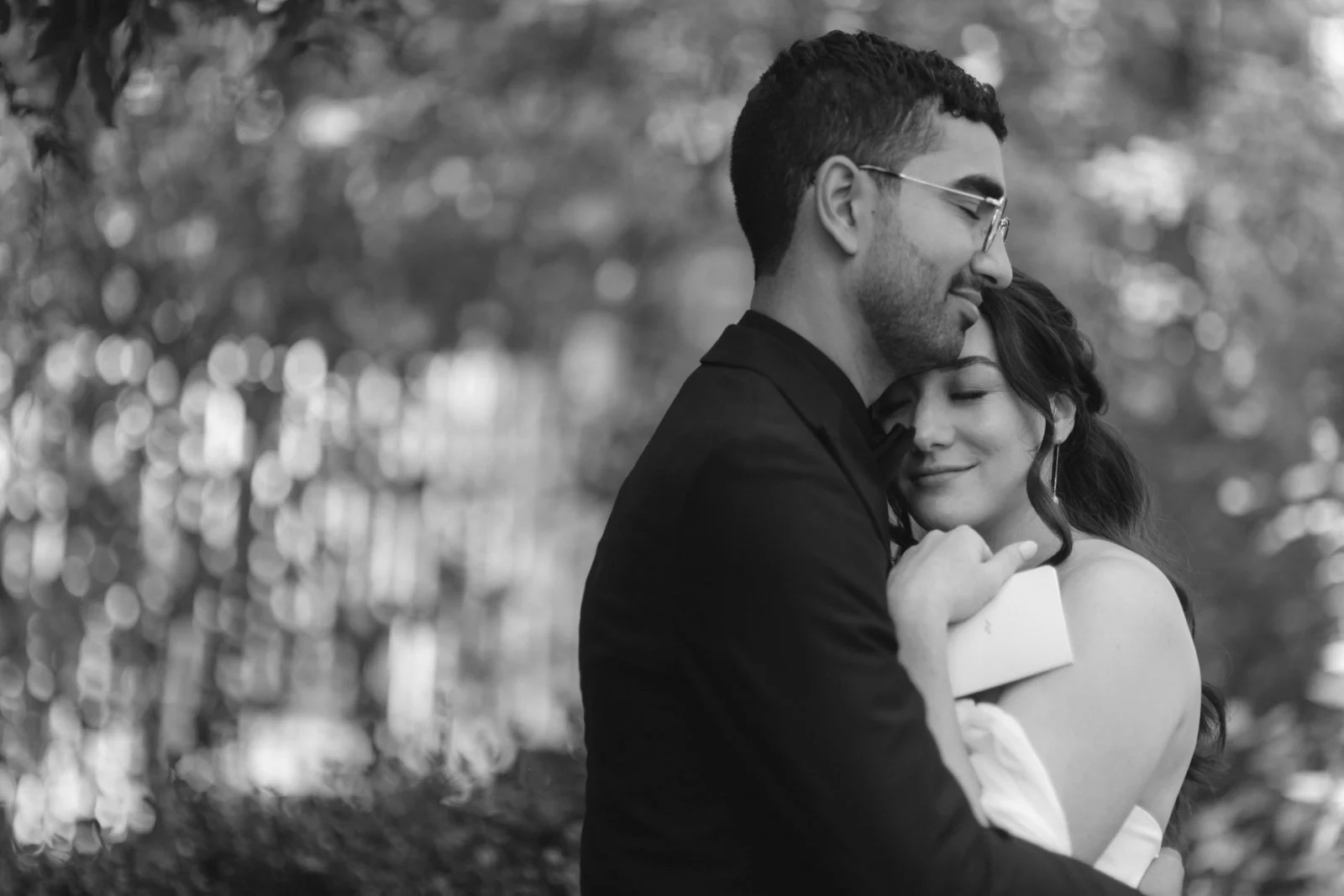 A black and white photo of a happy couple hugging outdoors, with the man in glasses and a dark jacket, and the woman with dark hair and a strapless dress, both smiling with eyes closed.