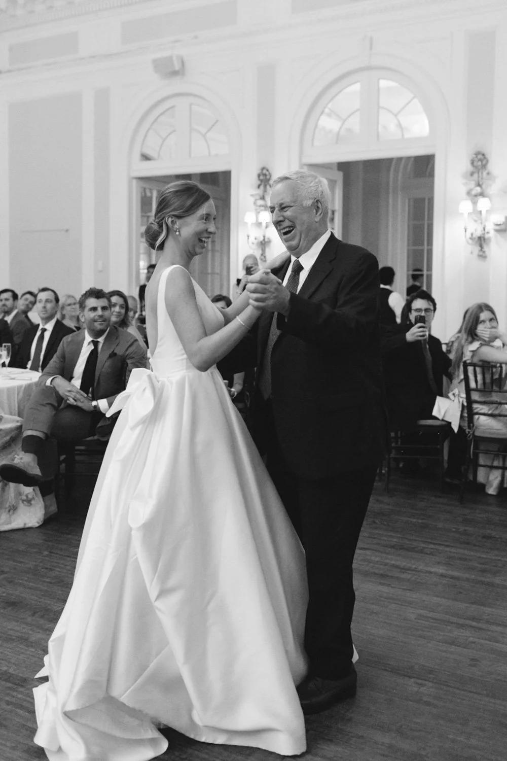 A bride in a wedding dress and an older man dance happily at a wedding reception, with guests seated and watching in a formal decorated hall.