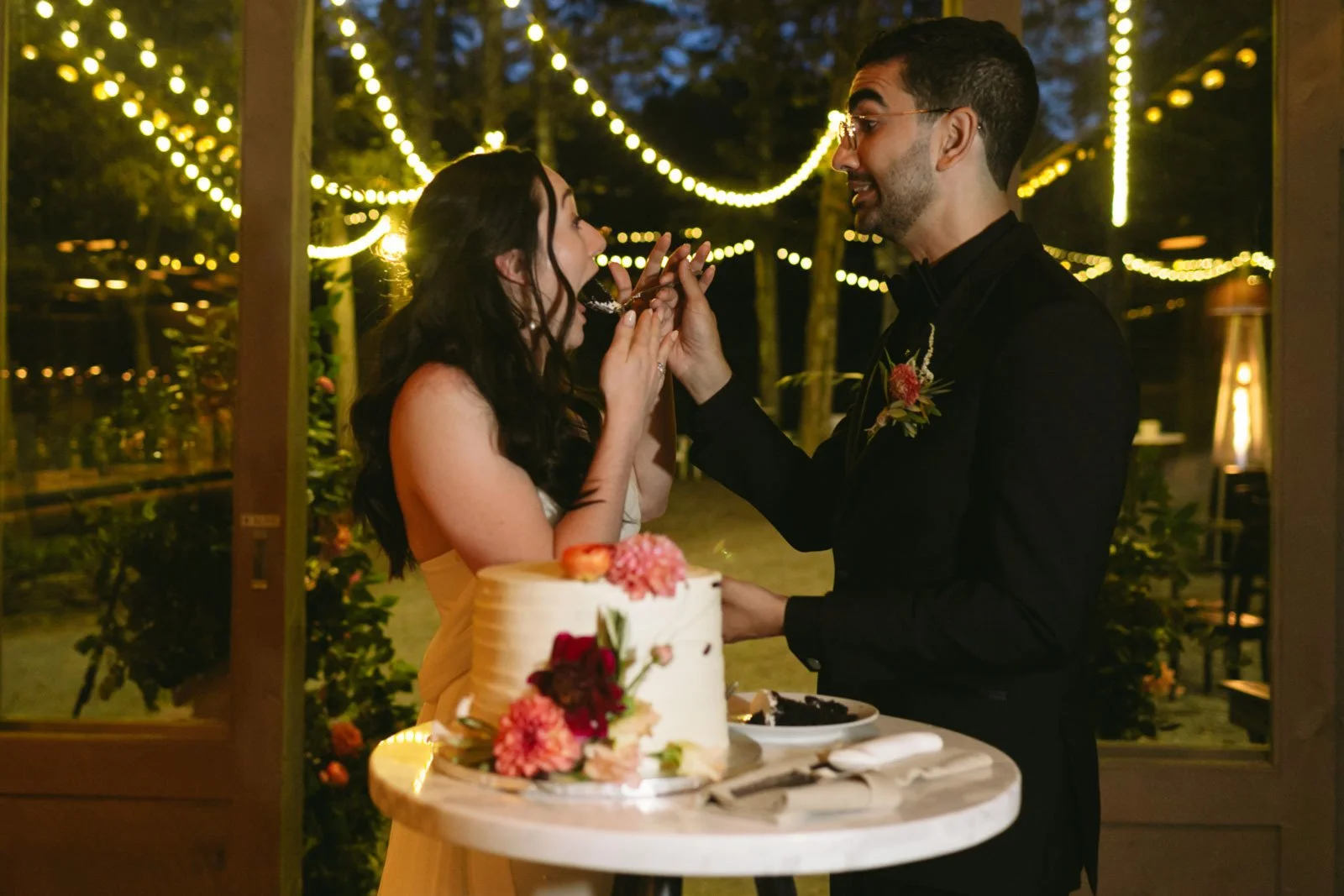 A couple at their wedding reception, with the bride in a yellow dress and the groom in a black tuxedo, sharing a cake-cutting moment with a cake decorated with flowers, under string lights in a cozy outdoor setting at night.