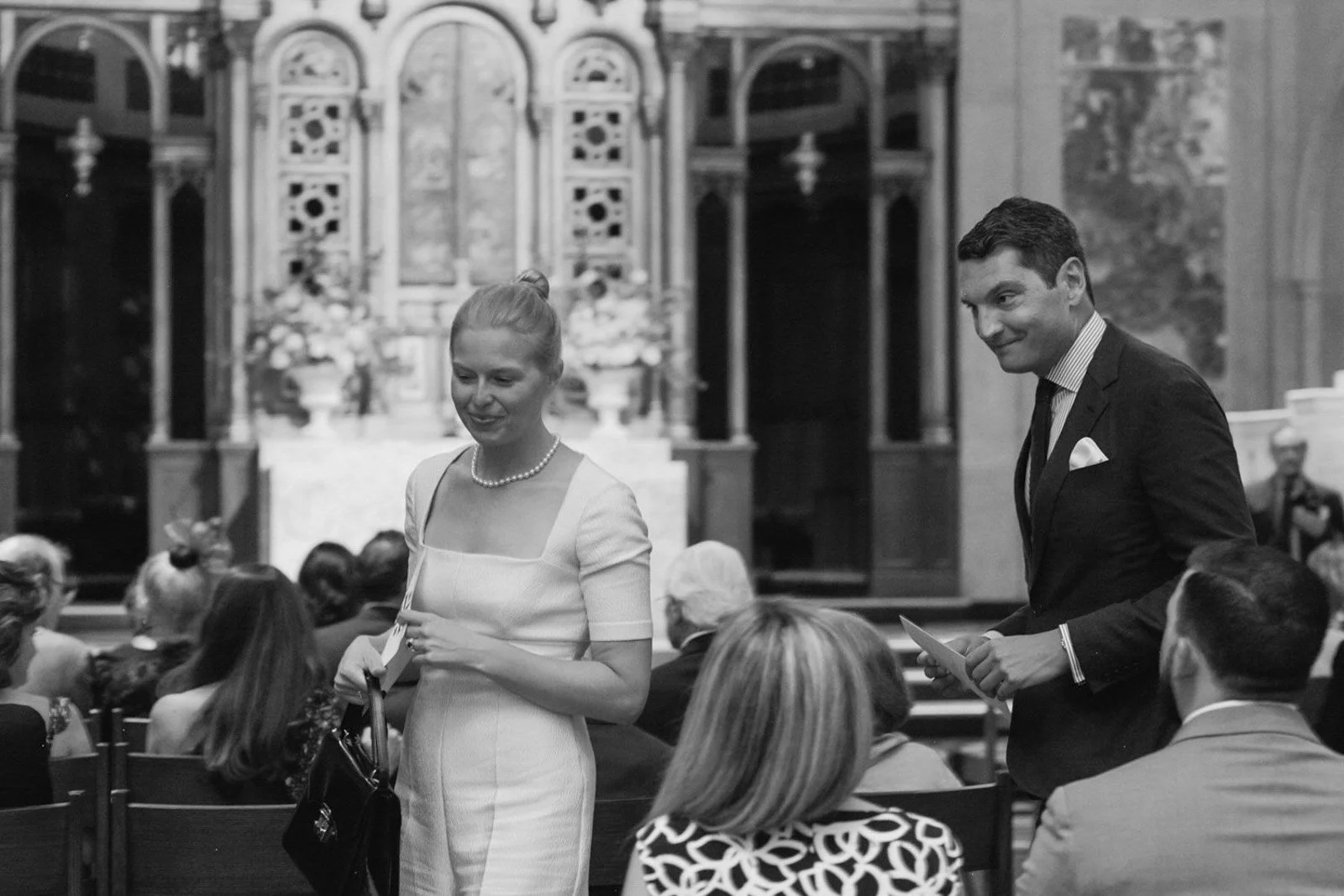 A black-and-white photo of a woman in a white dress and a man in a suit at a formal event, with several seated guests in the background and ornate architectural details behind them.