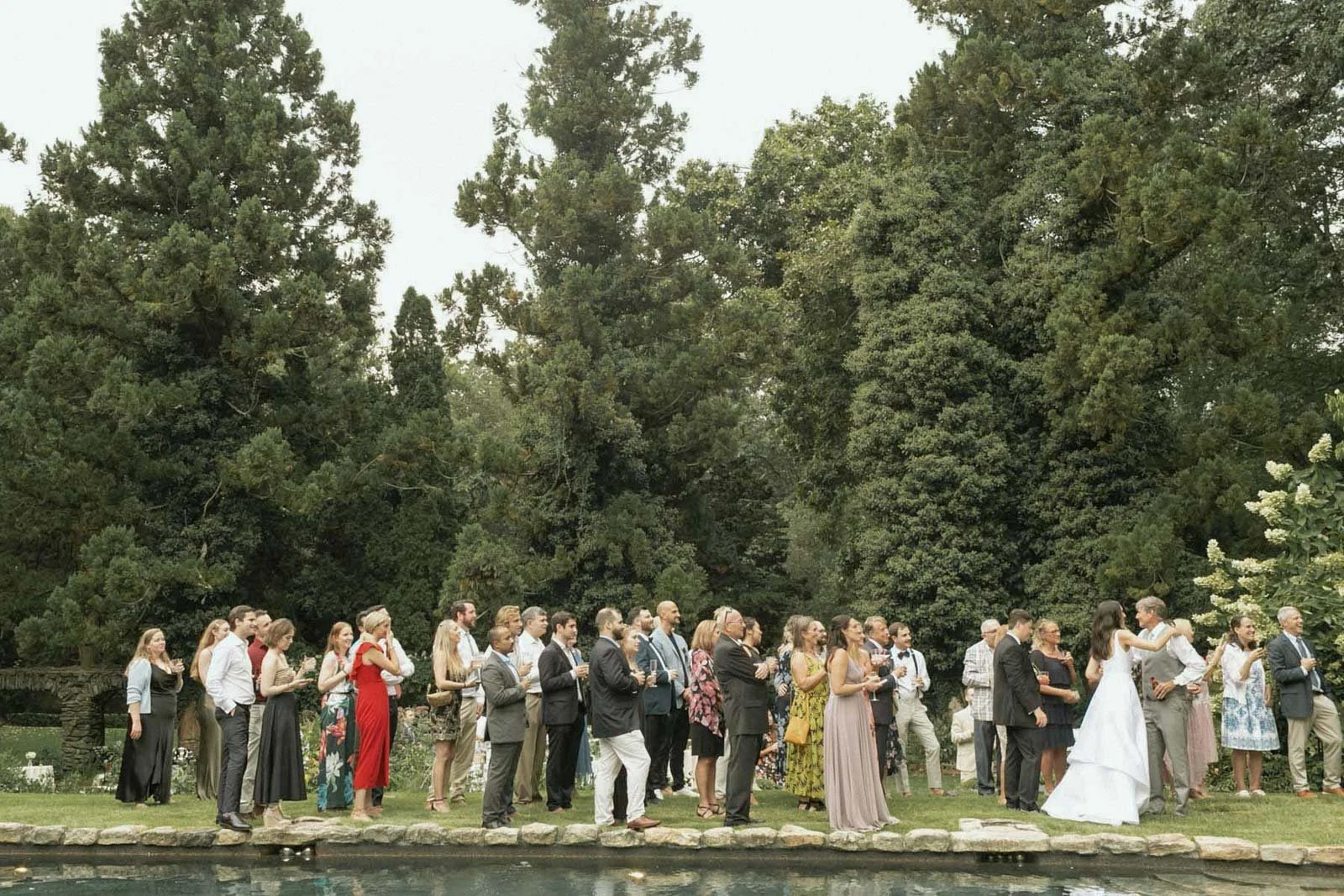 A group of people dressed in formal attire standing outdoors on a lawn, likely for a wedding, with tall trees in the background.
