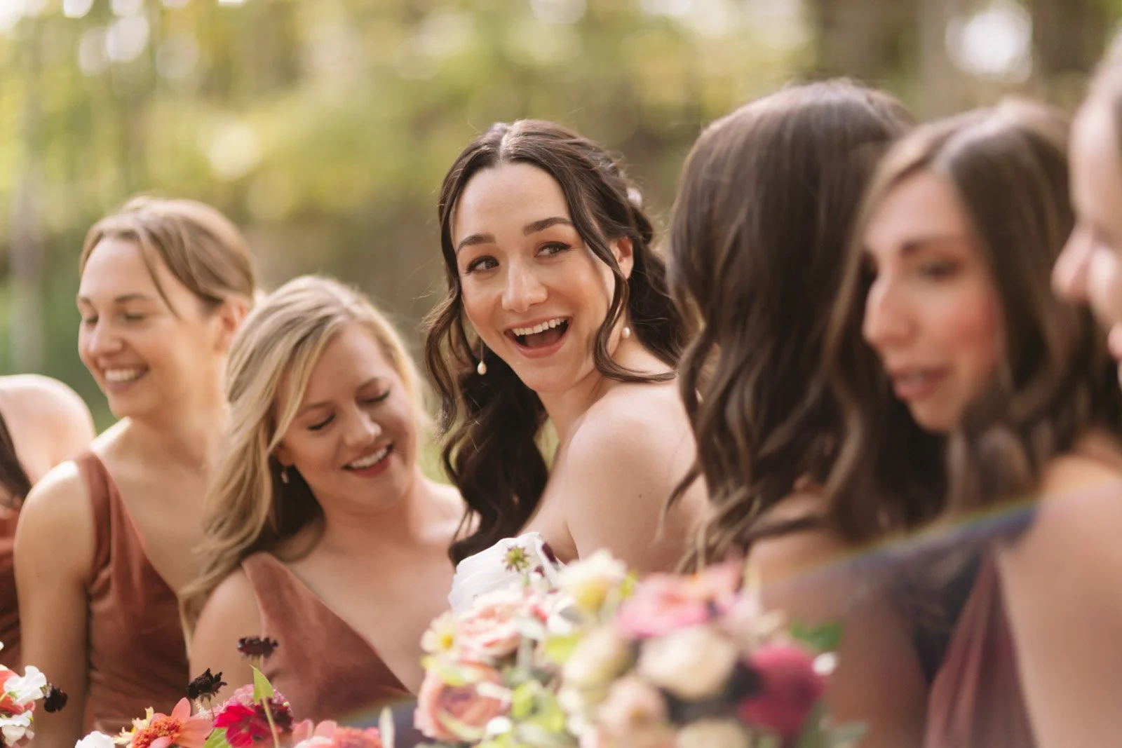 Group of women dressed in formal attire, smiling and engaging with each other outdoors during daytime, surrounded by flowers and greenery.