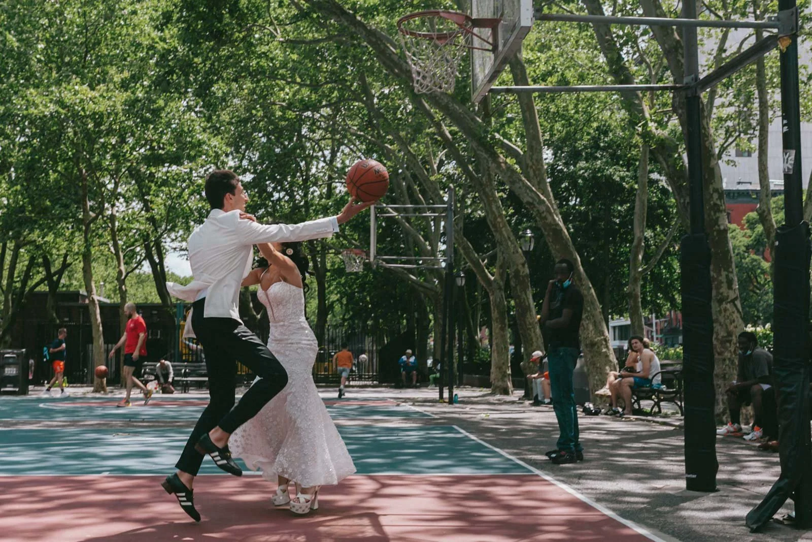A couple dressed in wedding attire playing basketball on an outdoor court with trees in the background.