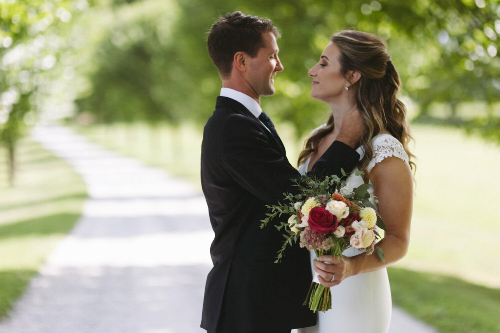 A bride and groom stand close together outdoors on a pathway, smiling. The bride holds a bouquet of red, white, and pink flowers with greenery, and they face each other affectionately in a lush green setting.
