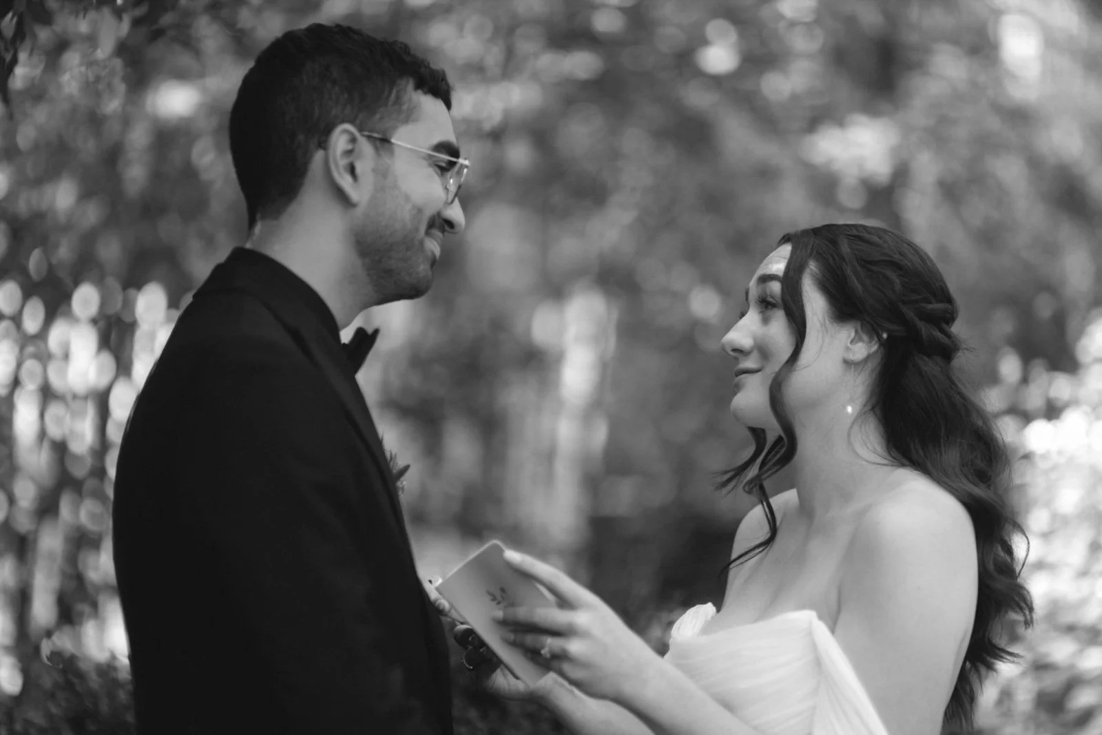 A black and white photo of a bride and groom standing outdoors, looking at each other, with the bride holding a small booklet or card.