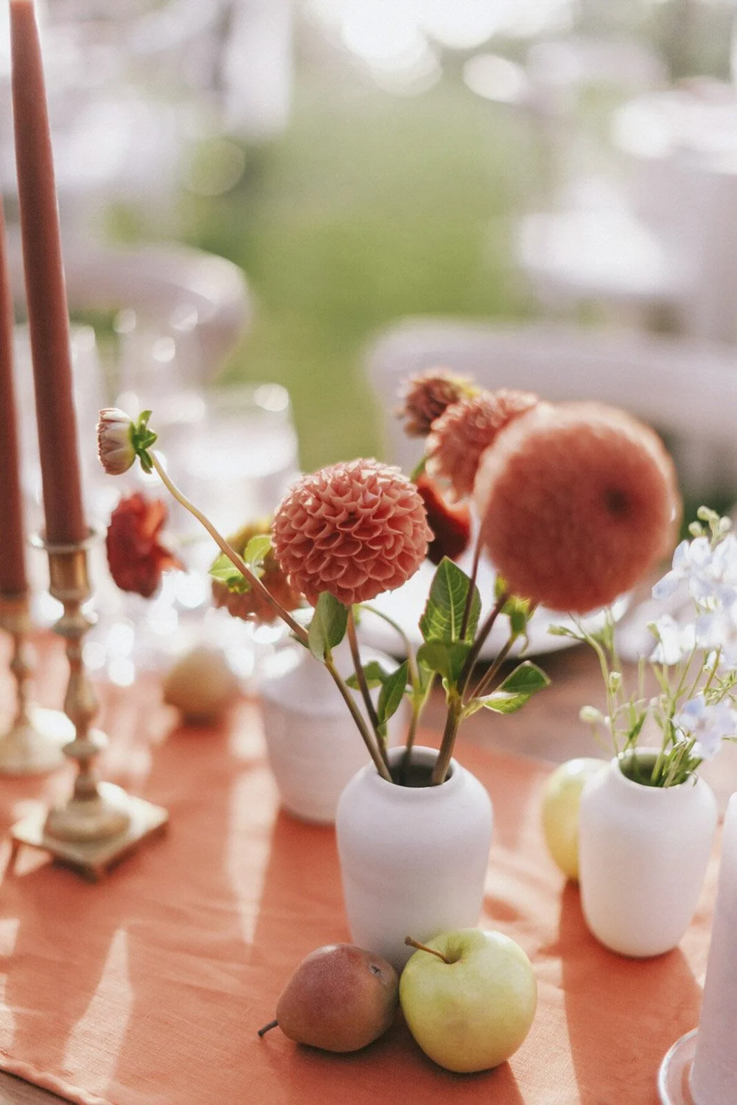 A bouquet of pink and peach dahlias in a white vase, placed on a wooden table with apples and a pear, surrounded by other small vases and decorative items, with a blurred background of greenery.