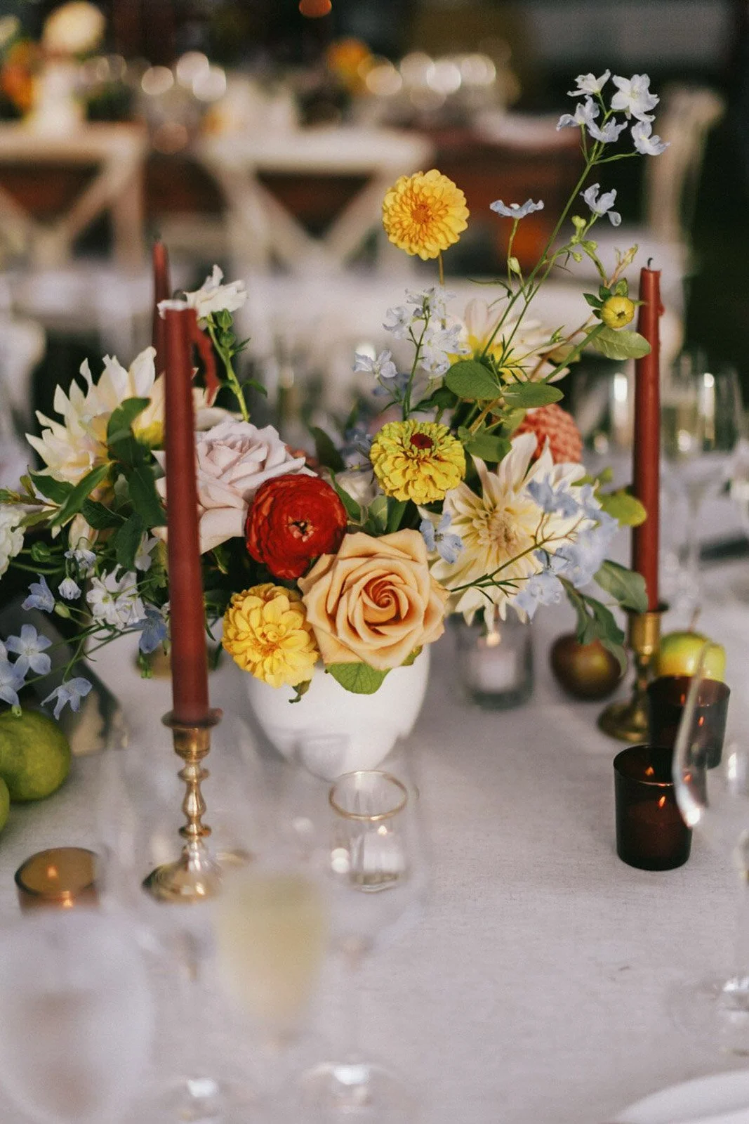 A floral centerpiece with peach roses, yellow and red flowers, and white and blue blooms on a white tablecloth surrounded by candles and tableware.