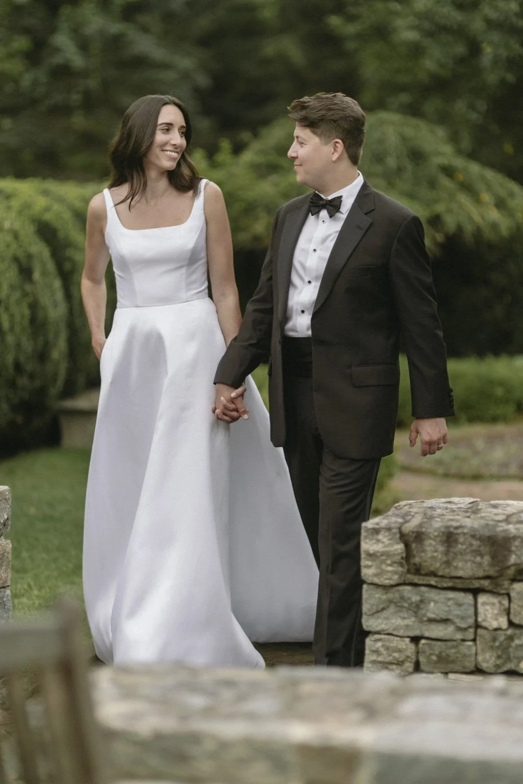 A bride and groom holding hands outdoors in a garden, smiling at each other, the bride in a white dress and the groom in a black tuxedo.