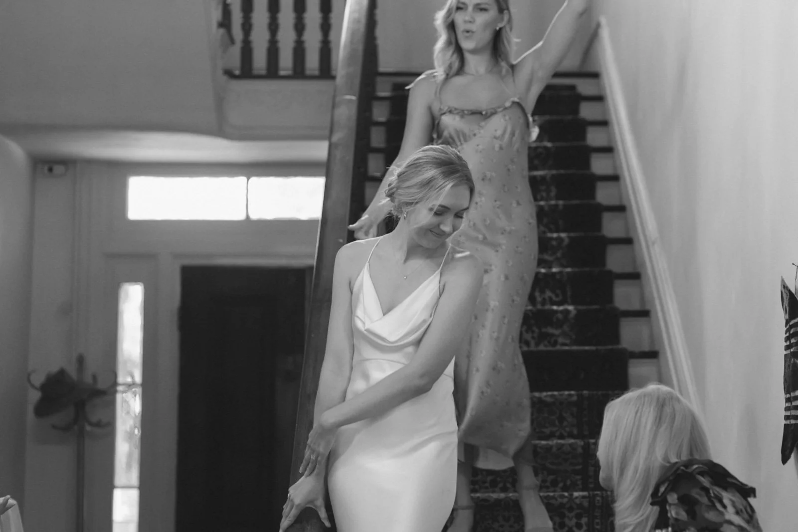 Two women descending a staircase, one in a light-colored slip dress looking down and smiling, the other in a patterned dress with a surprised or playful expression, with a third woman crouched at the bottom of the staircase.