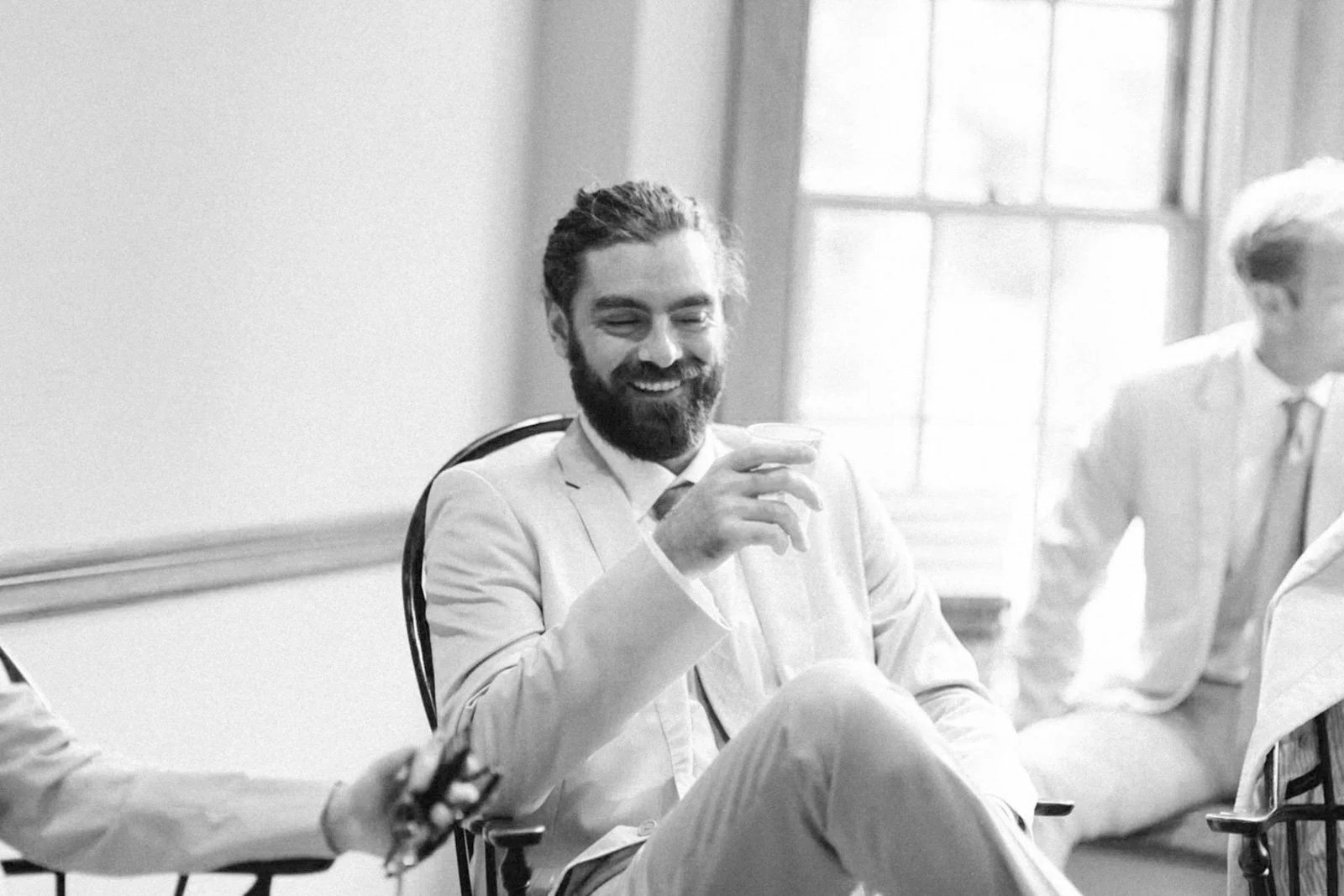 Man with a beard and smiling, wearing a light-colored suit, sitting in a meeting room, holding a cup, with windows in the background.