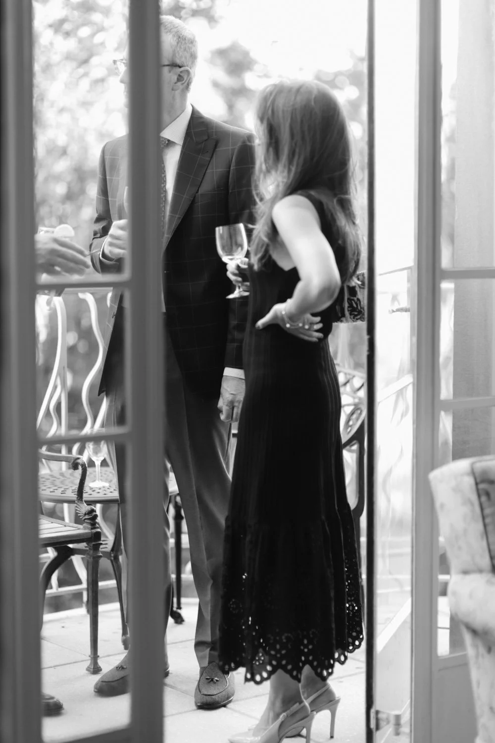 People at an indoor social gathering, holding wine glasses, viewed through glass doors.