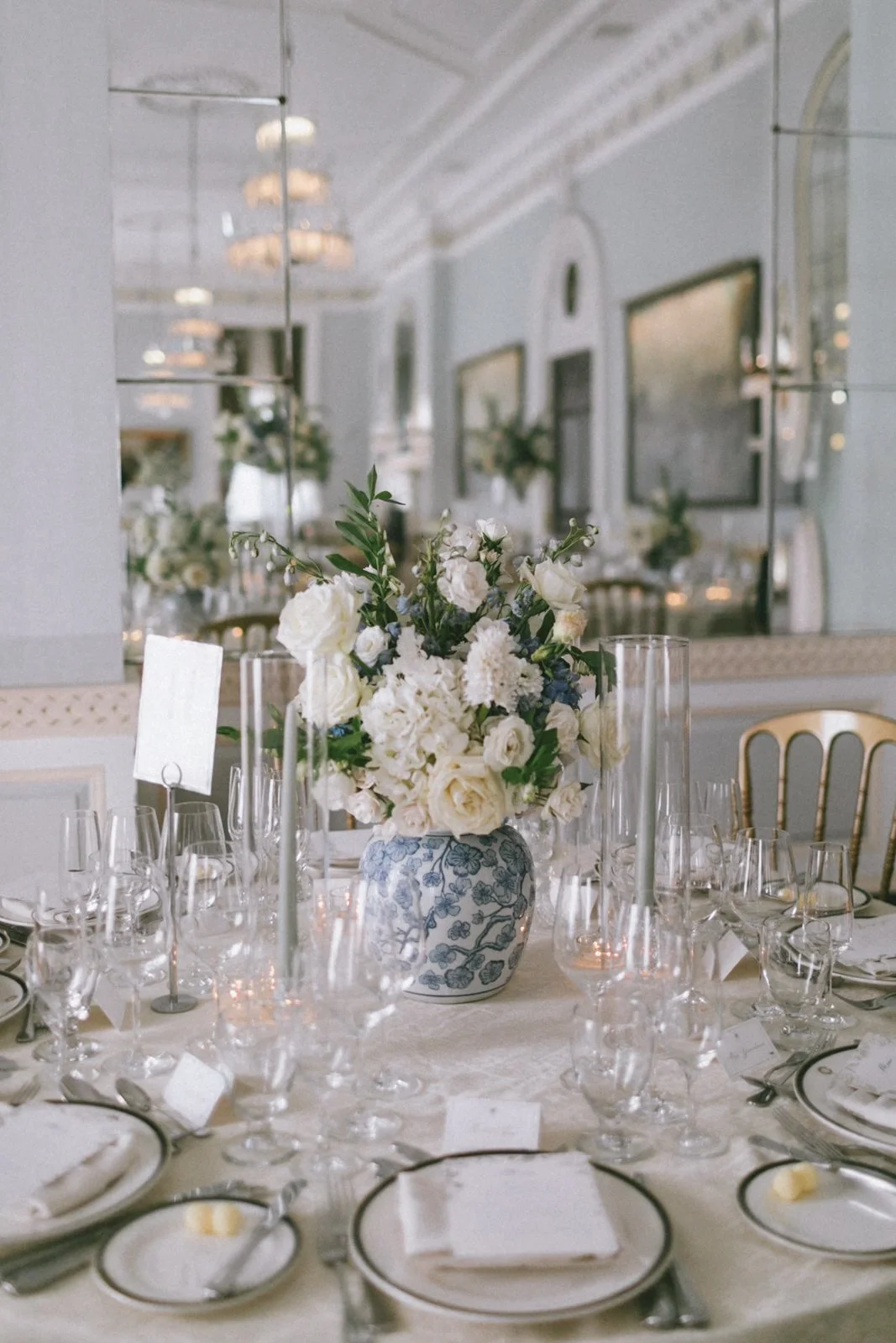 A floral centerpiece with white roses and greenery on a decorated table set for a formal event in an elegant room with chandeliers and mirrors.