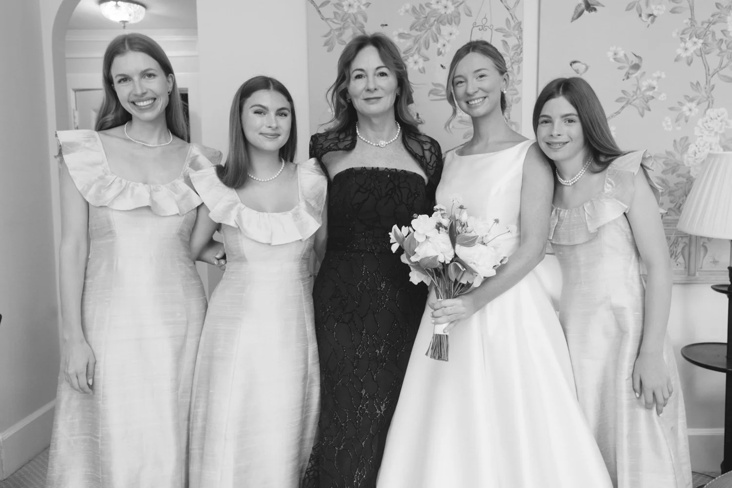 A bride in a wedding dress holding a bouquet of flowers standing next to five women in matching elegant dresses, with one woman in the center wearing a black dress, in a decorated room.
