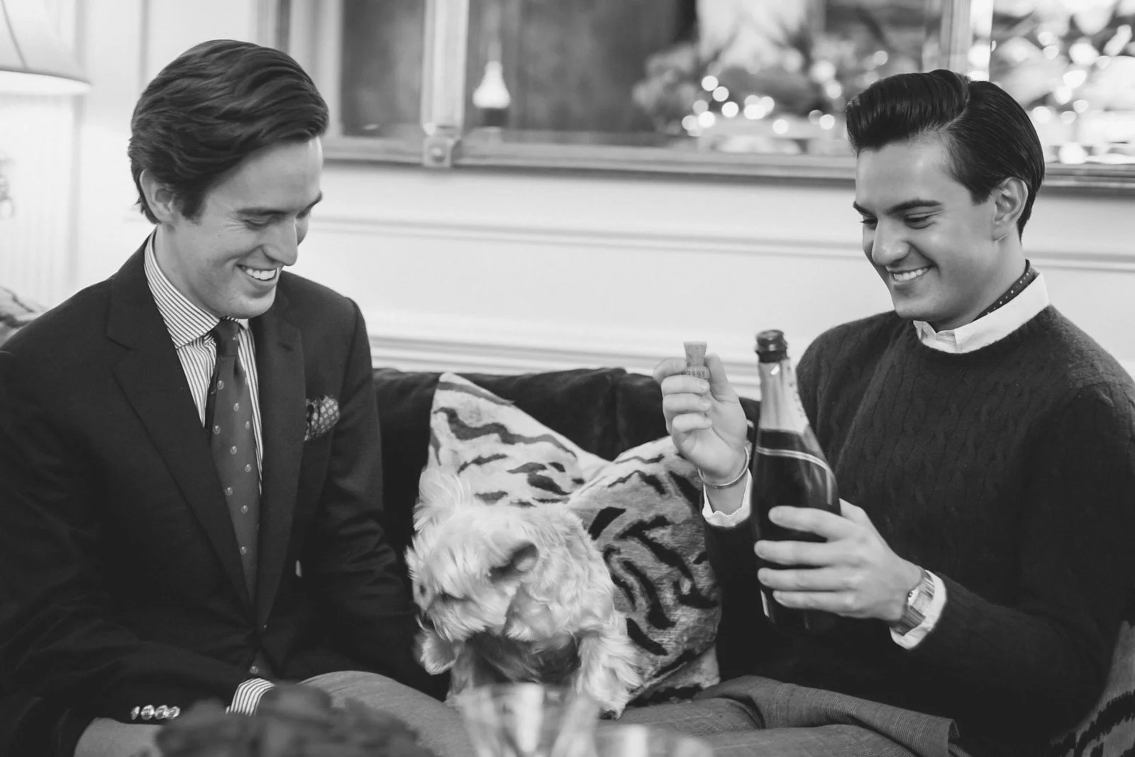 Two men dressed in business and casual attire sitting on a couch with a small dog between them, smiling and holding a champagne cork and bottle, in a cozy indoor setting.