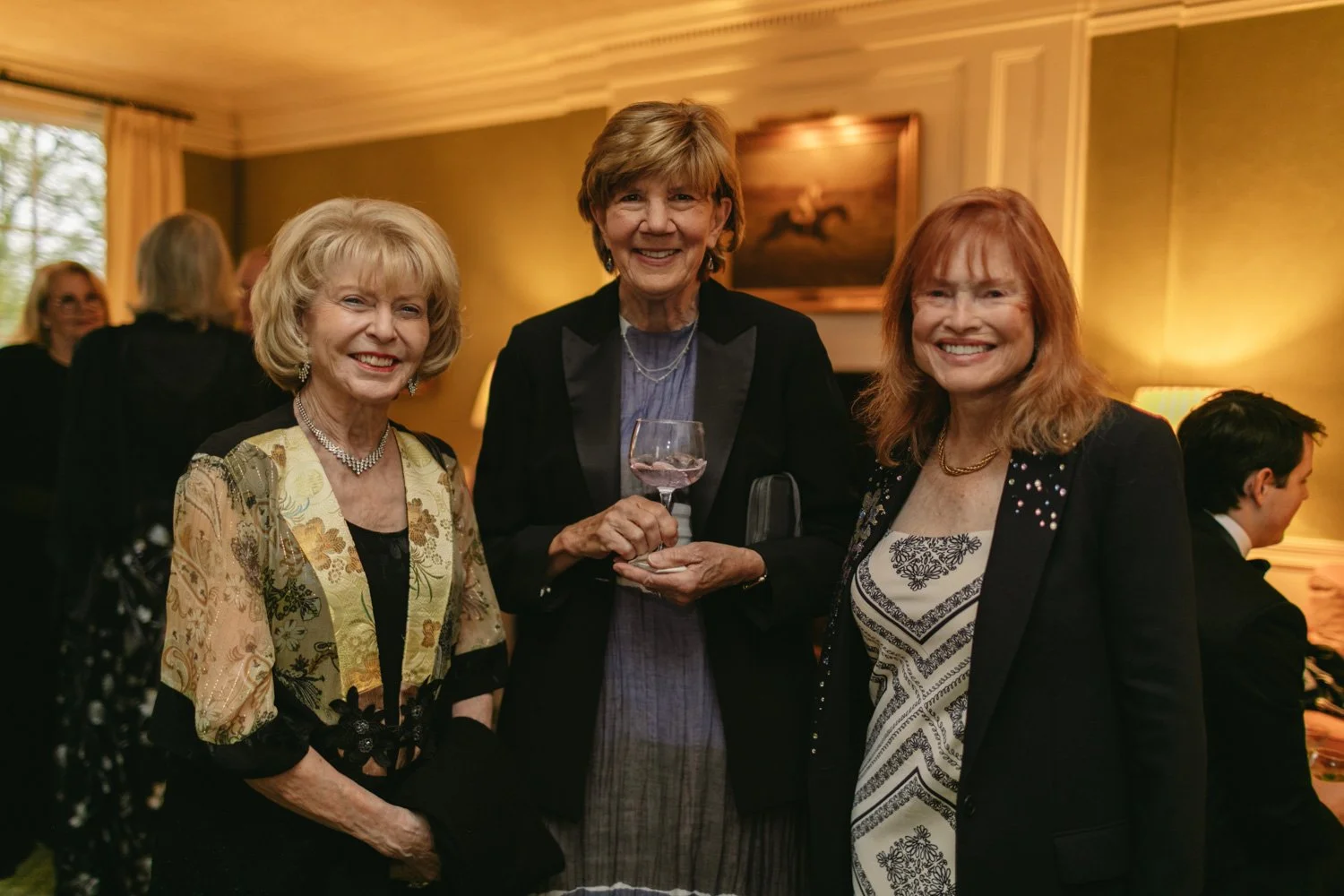 Three women smiling at a social gathering, with one holding a glass of wine, in a warmly lit room with wooden walls and a painting in the background.