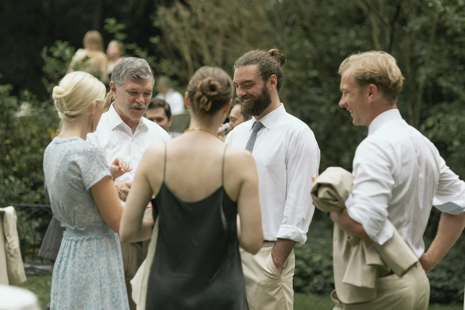People dressed in formal attire gathered outdoors, engaging in conversation at a social event in a garden setting.