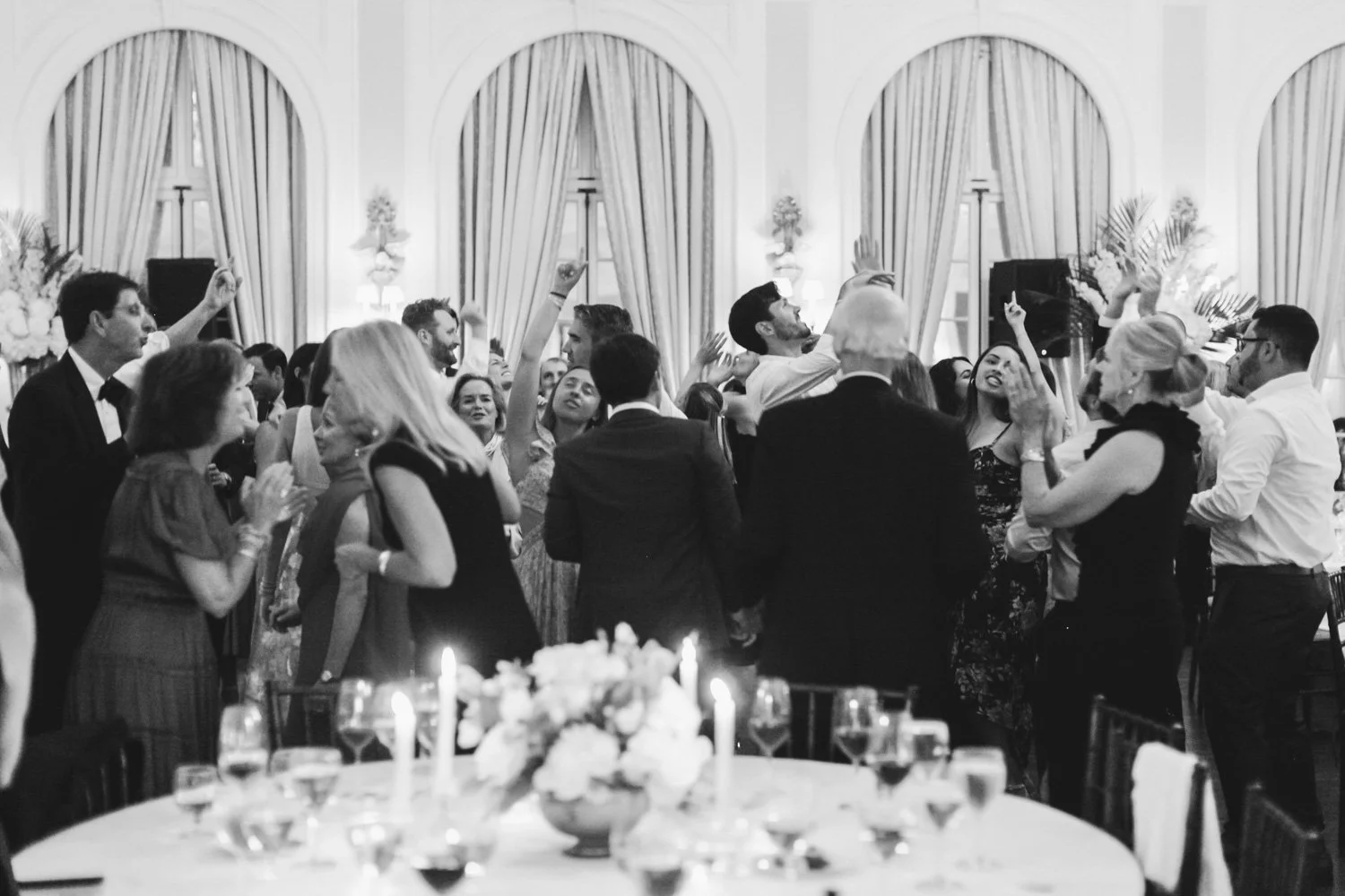 People dancing and celebrating at a formal indoor event, with elegant curtains and floral decorations in the background.