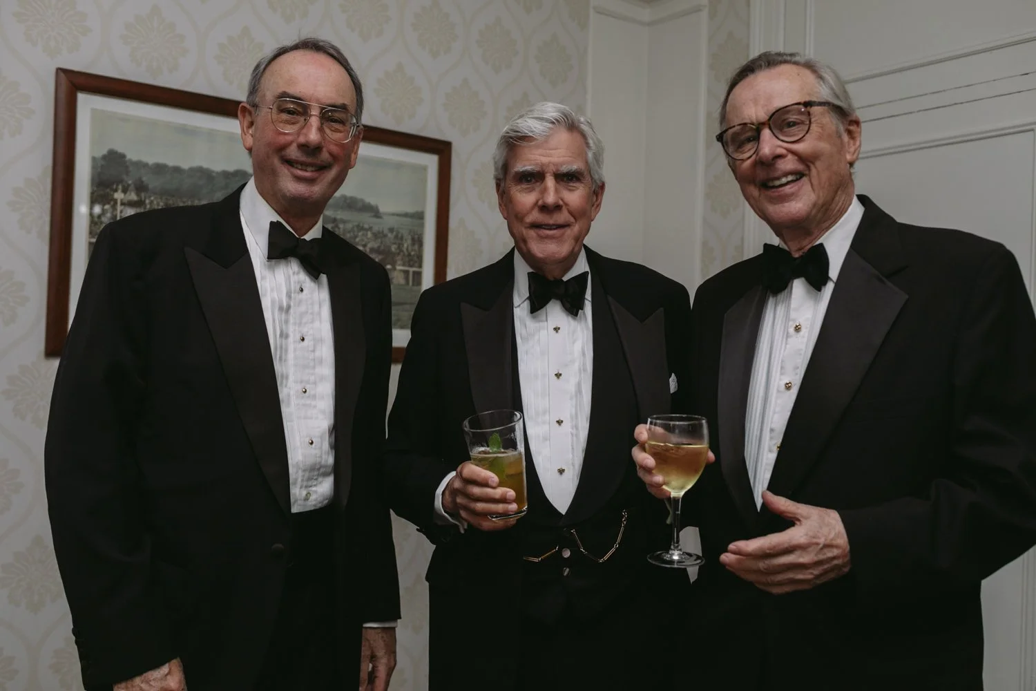 Three elderly men in tuxedos and bow ties holding drinks at a formal gathering.