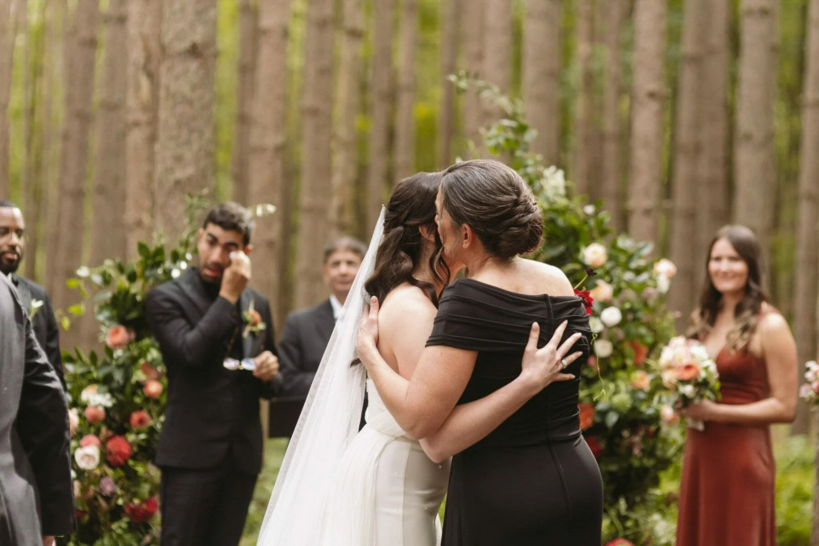 Two women sharing a kiss at a wedding ceremony outdoors, surrounded by friends and floral decorations in a wooded area.