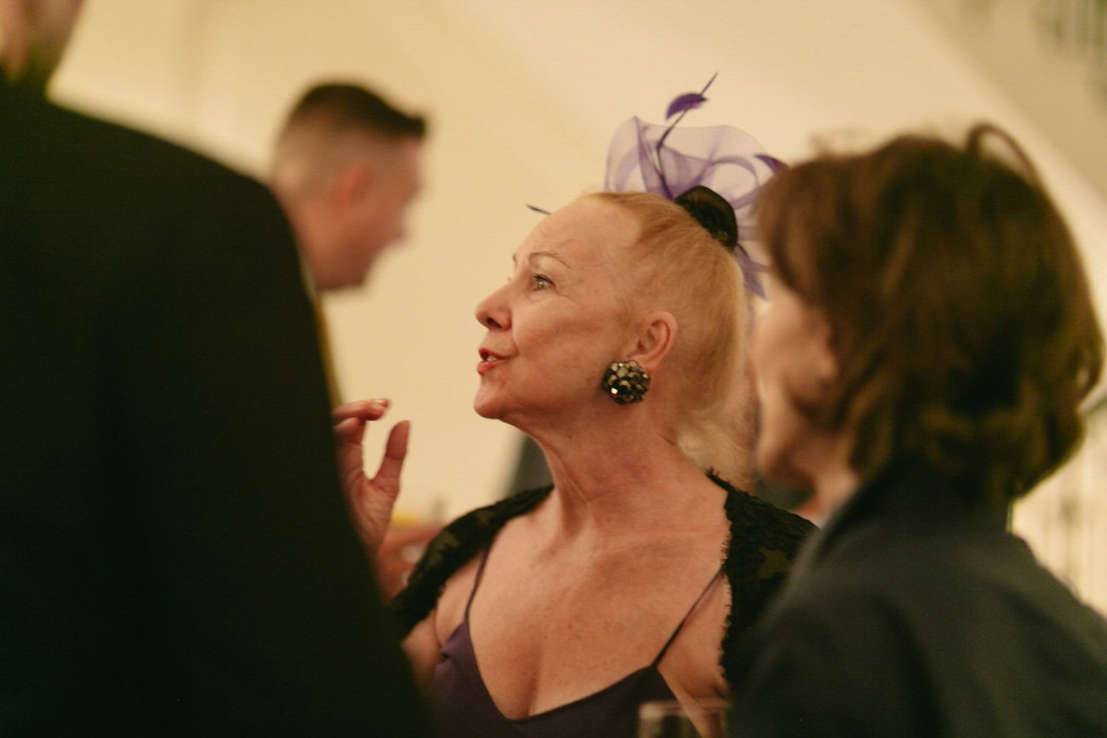 An older woman with blonde hair, wearing earrings, a purple fascinator, and a black lace top, engaged in conversation with a man and a woman at a social event.