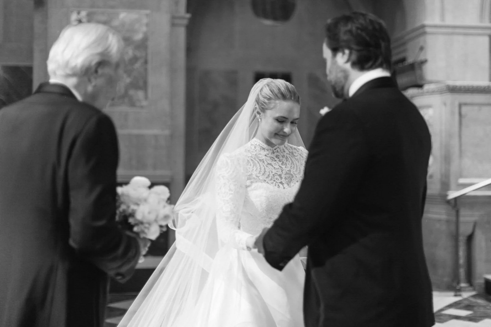 A bride and groom holding hands during a wedding ceremony in a church, with an officiant standing nearby holding a bouquet of flowers.