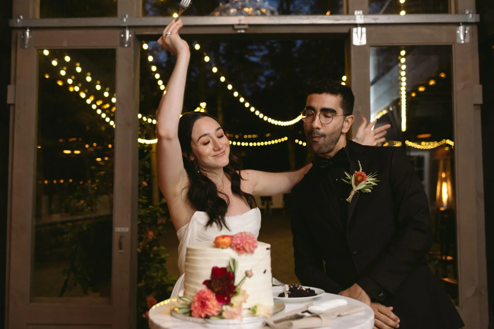 A bride and groom celebrating at their wedding reception, standing behind a cake with flowers, with string lights hanging in the background during the evening.