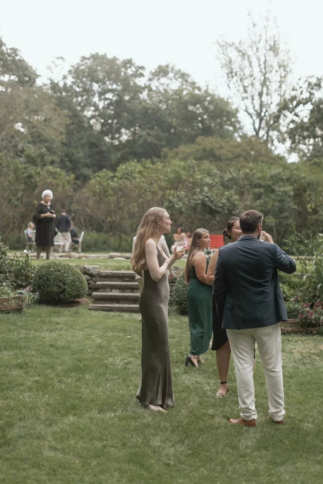 Group of people dressed in formal attire socializing outdoors in a garden setting with trees and shrubs, during daytime.