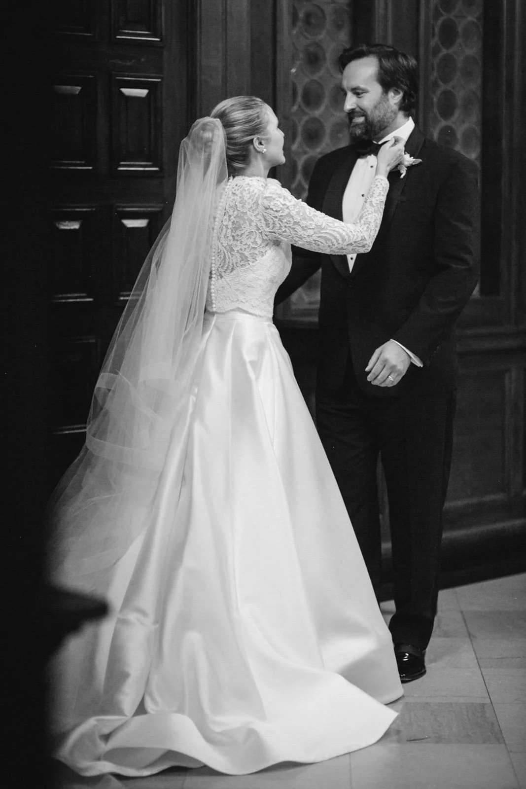 A bride and groom sharing a dance during their wedding in a formal setting, with the bride in a lace wedding dress and veil, and the groom in a black tuxedo with a bow tie.