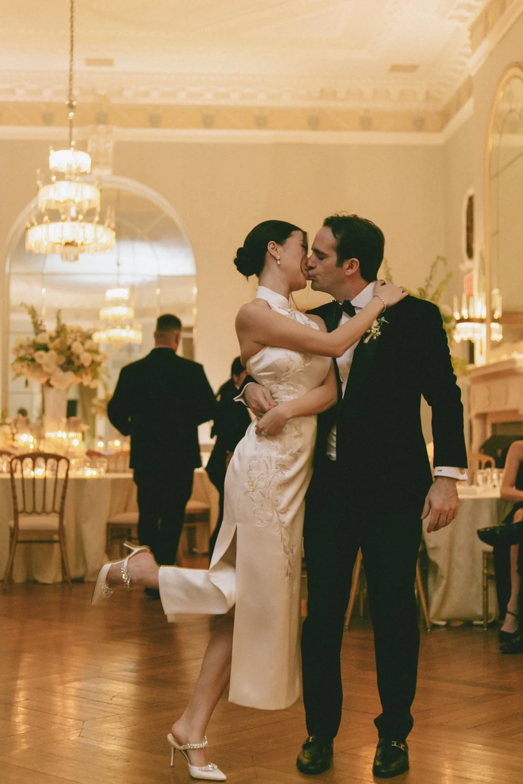 A couple dressed in wedding attire sharing a kiss and dance at their wedding reception in an elegant ballroom with chandeliers and floral decorations.