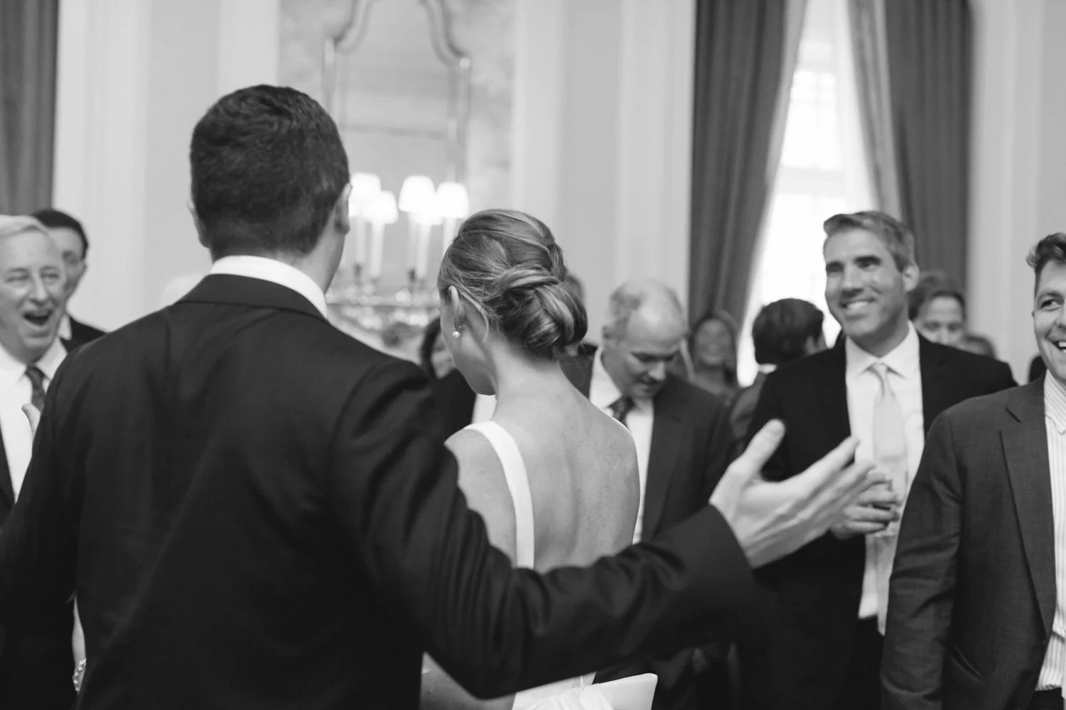 A black-and-white photo of a wedding reception with guests smiling and talking in a decorated, elegant room with high ceilings and large windows. The bride is facing away from the camera, with an updo hairstyle, and the groom is partially visible fro