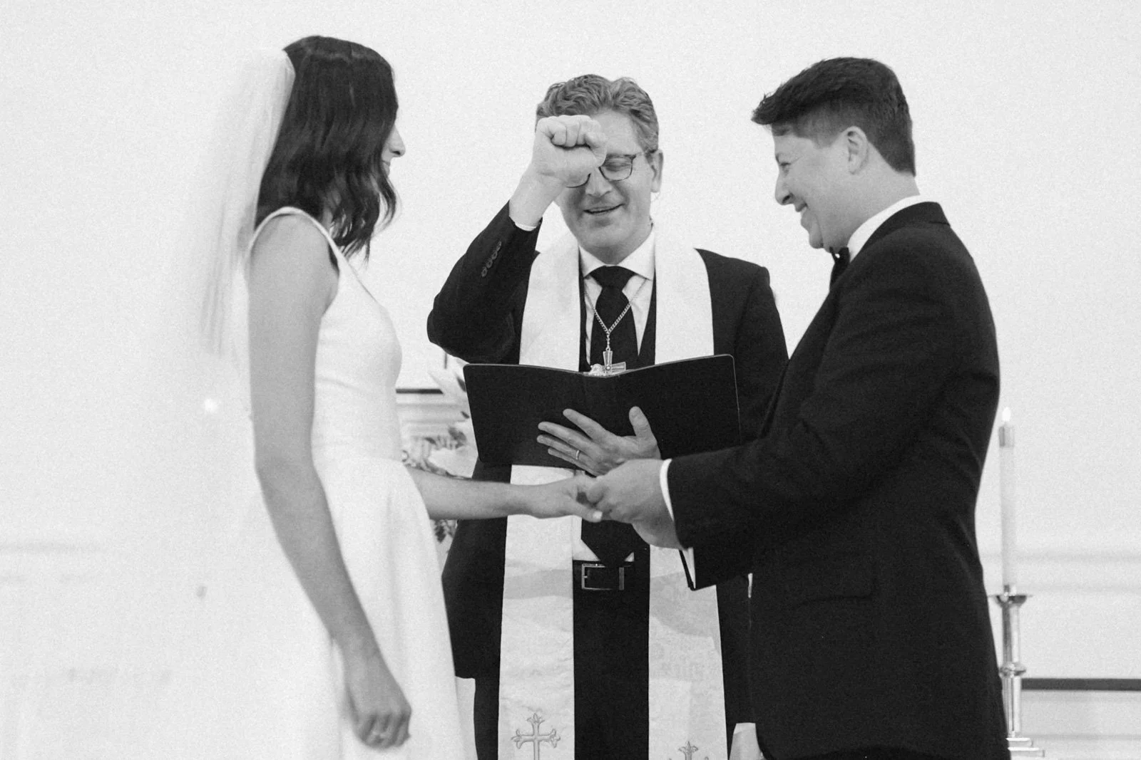 A couple getting married during a wedding ceremony, holding hands, with an officiant reading from a book.