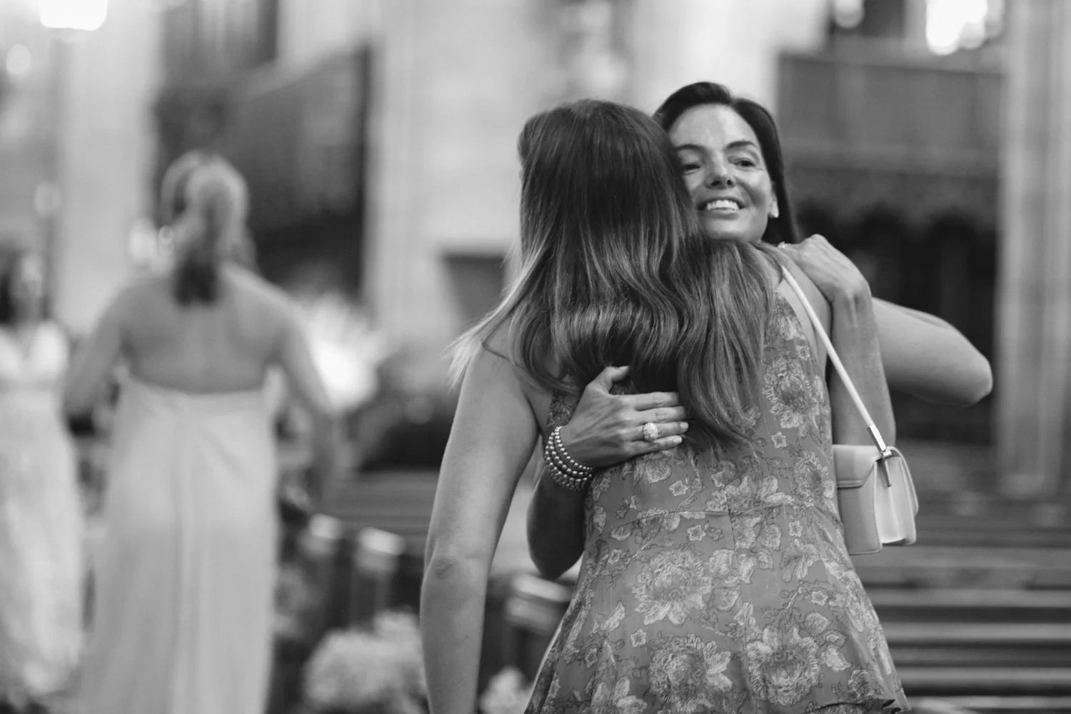 Two women hugging and smiling at each other inside a church with other people in the background.