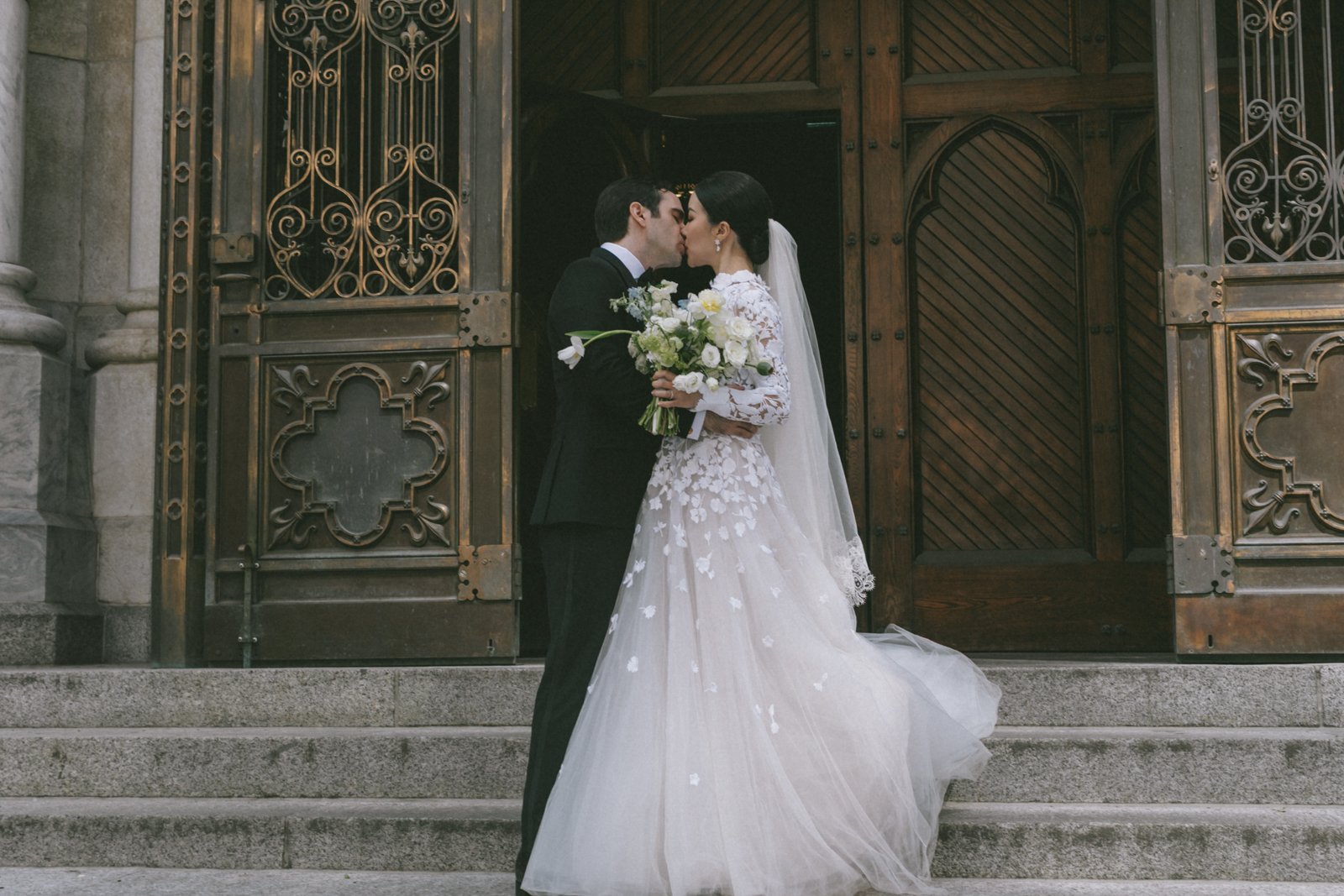 A bride and groom kiss on the steps of a church. The bride wears a white lace wedding dress and veil, and holds a bouquet of white flowers. The groom wears a black tuxedo. They stand in front of an ornate wooden and metal church door.