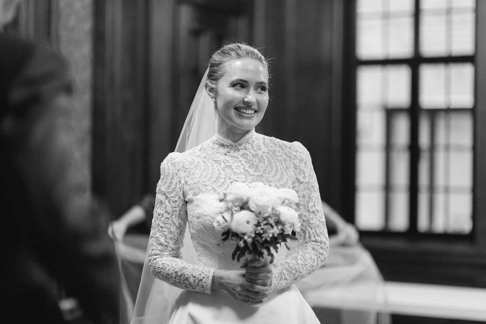 A bride in a lace wedding dress holding a bouquet of flowers, smiling in a room with large windows.