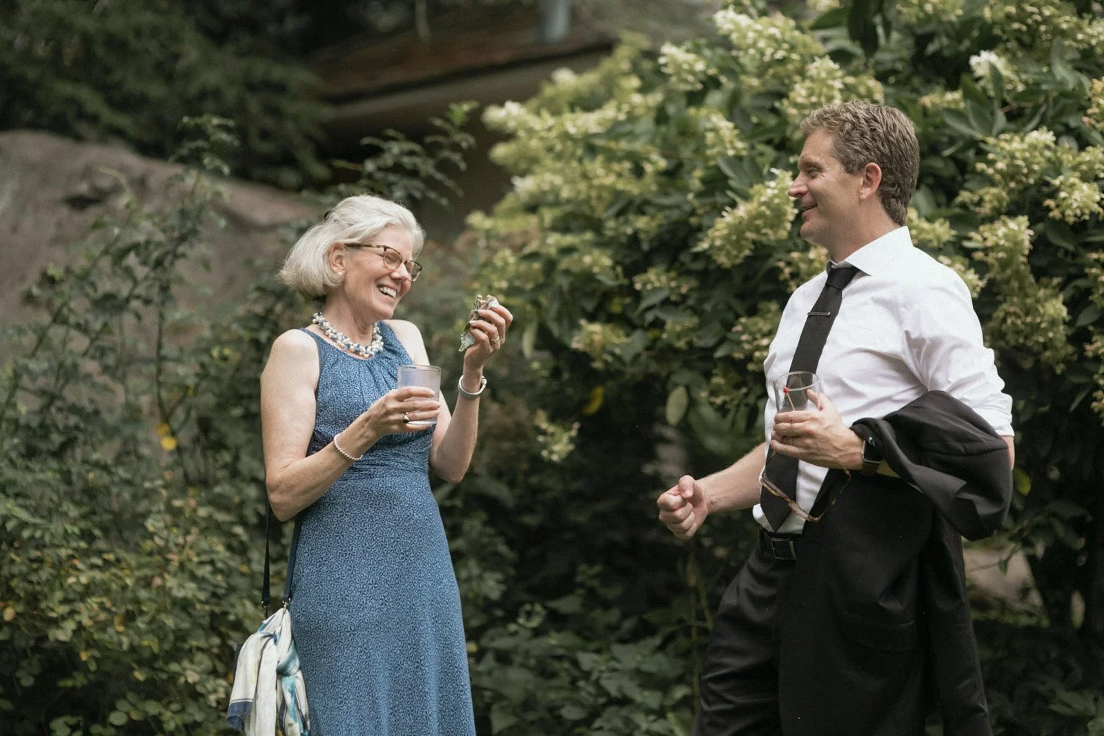 An elderly woman with glasses and white hair, wearing a blue dress and pearl necklace, laughing and holding a drink while talking to a man in a white shirt and black tie, who is also holding a drink, outdoors among greenery.