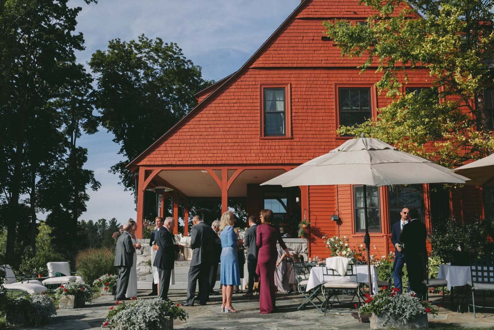 People gathered outside a red wooden house with umbrellas, dressed in formal attire, for a wedding at Court Hill in The Berkshires.