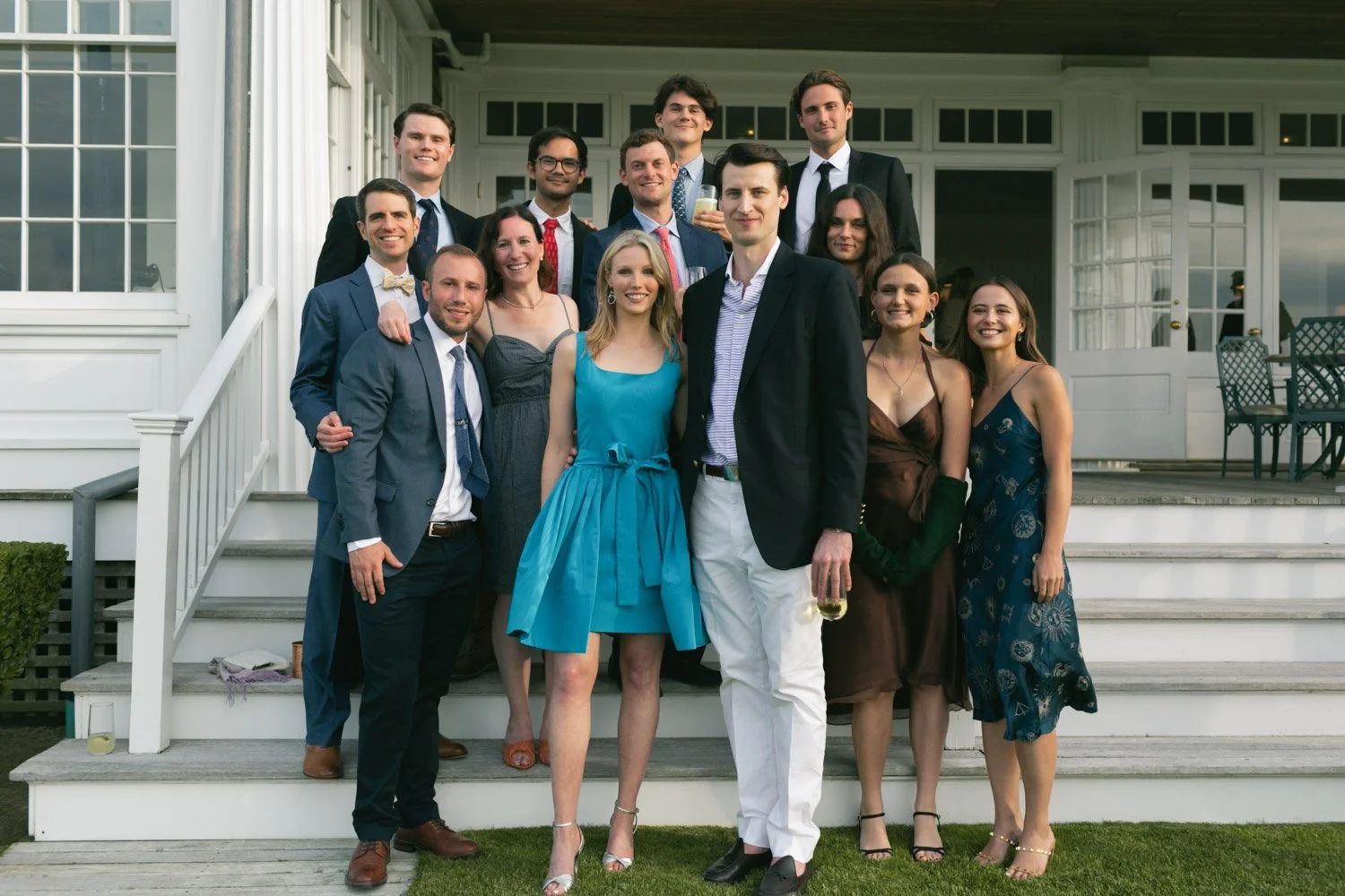 Group of young adults dressed in formal attire on the steps of a white house or building, smiling and holding drinks, celebrating an event or gathering.