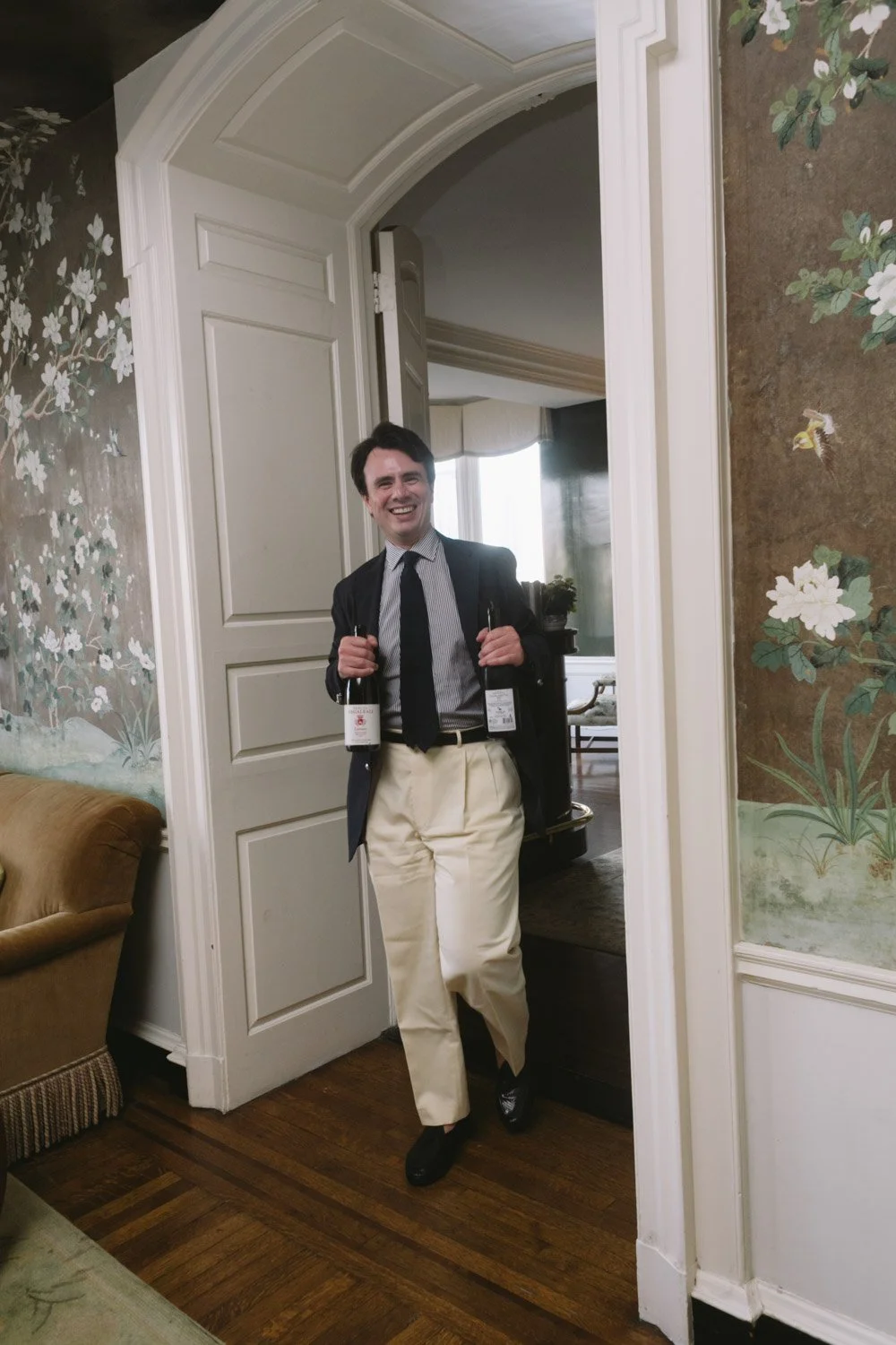 A man in dressy attire holding two bottles of wine indoors near a doorway, smiling.