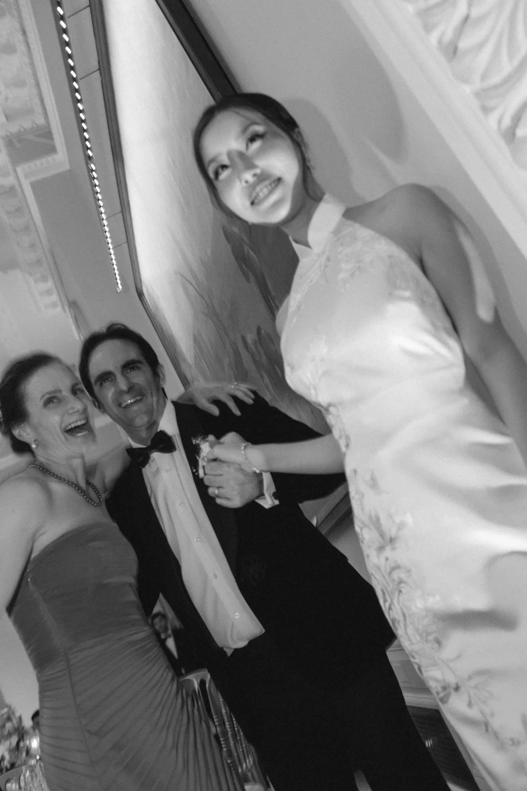 A black and white photo of three people dressed in formal attire at a wedding or formal event. A woman in a strapless gown is on the left, another woman in a gown with floral embroidery in the front is on the right, and a man in a tuxedo with a bow t