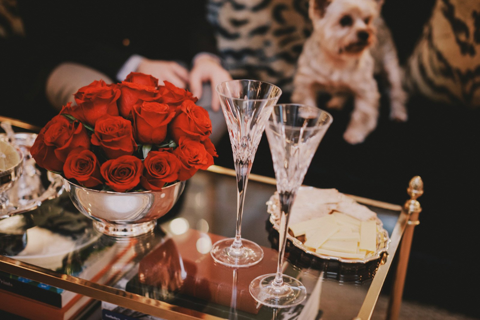 A silver bowl filled with red roses, two crystal champagne flutes, a silver tray with white chocolate pieces, and a set of small cream-colored figures on a glass table.