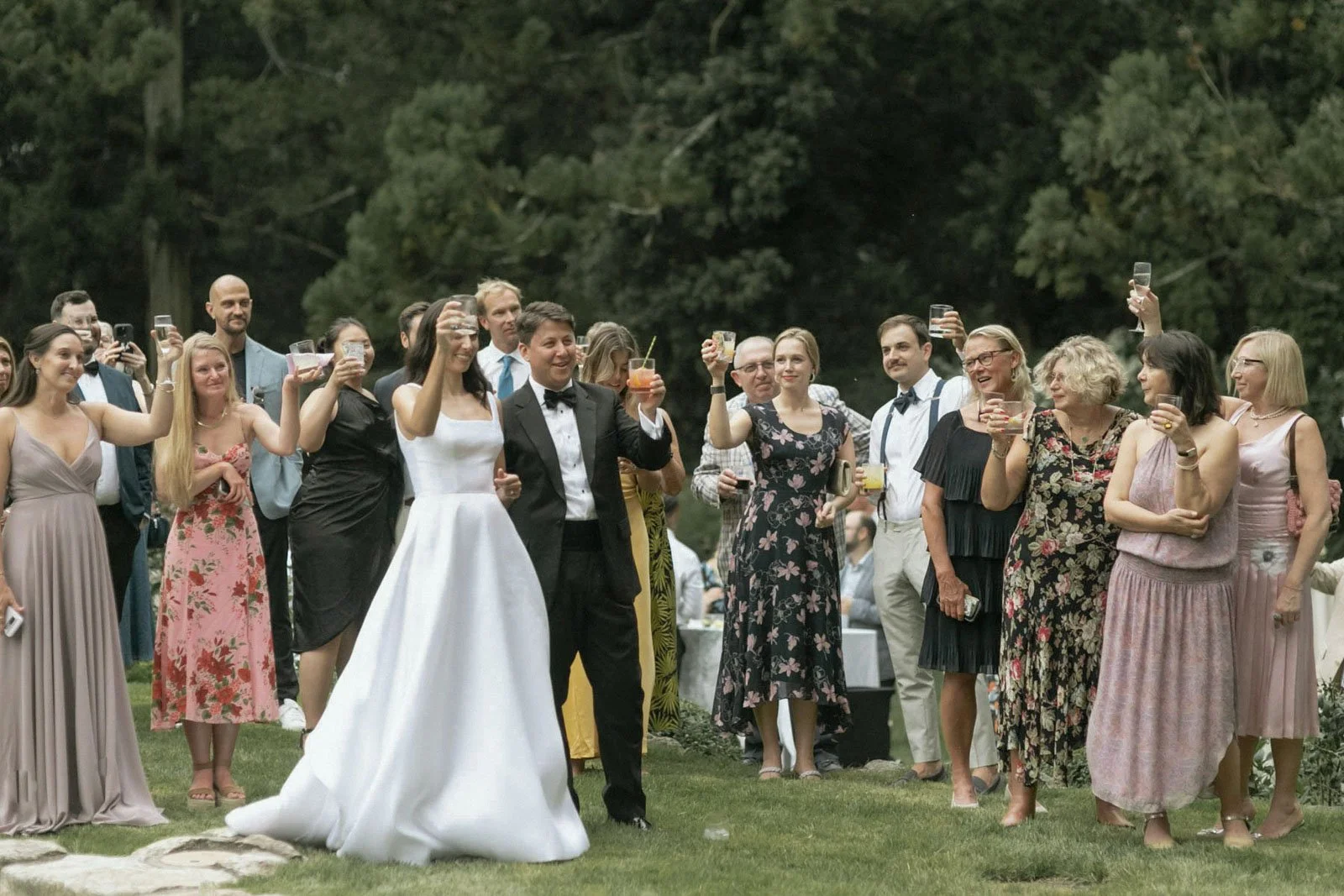 A group of people at a wedding reception outdoors, dressed in formal attire, raising their glasses in a toast, with trees in the background.