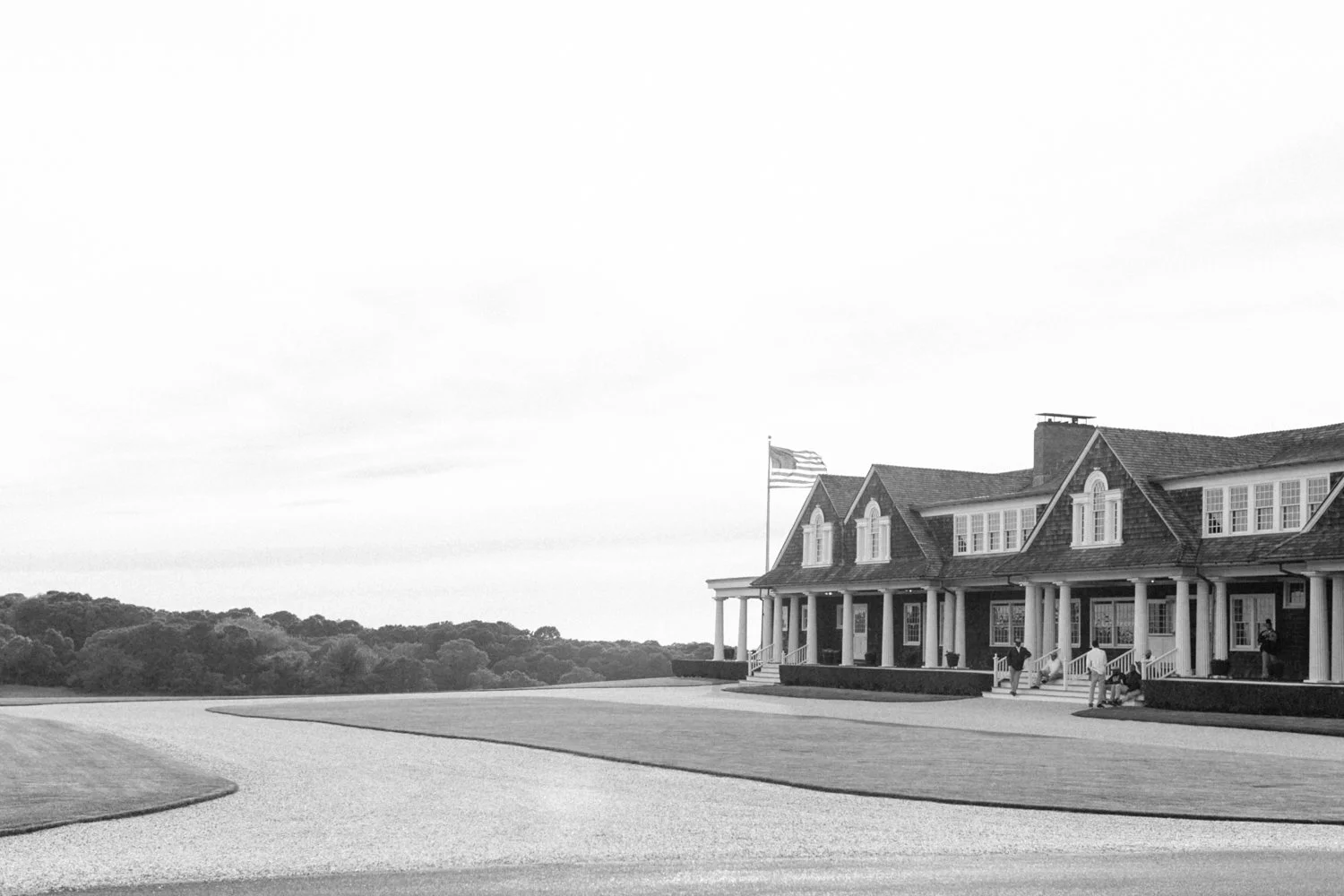 A large house with a porch, multiple windows, and an American flag flying on a pole beside it. Several people are gathered on the porch. The house is located in a spacious area with a curved driveway and trees in the background.
